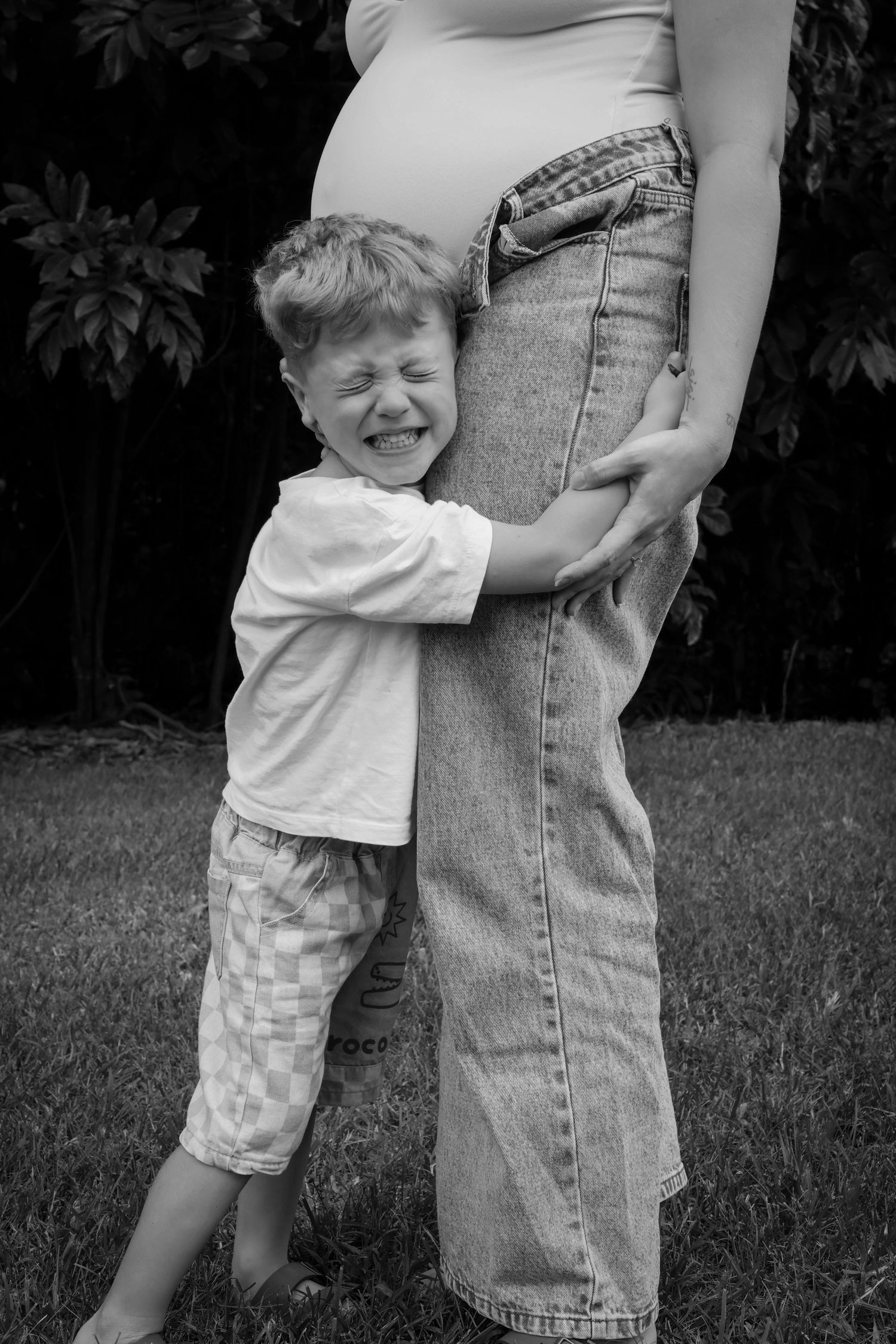 Black and white photo of a young boy hugging his pregnant mother's belly outdoors in a garden.