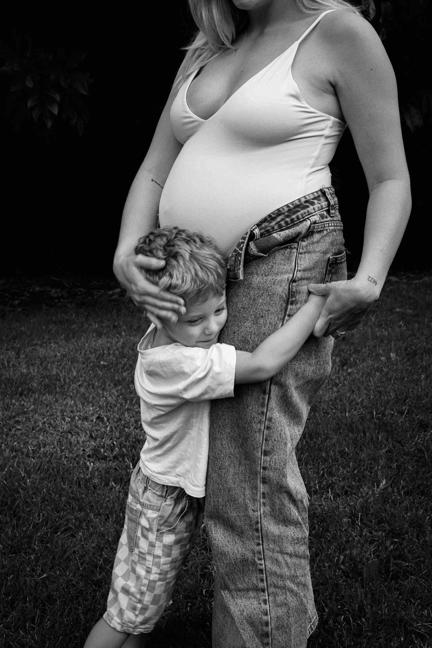 Black and white photo of a young boy hugging his pregnant mother's belly outdoors.