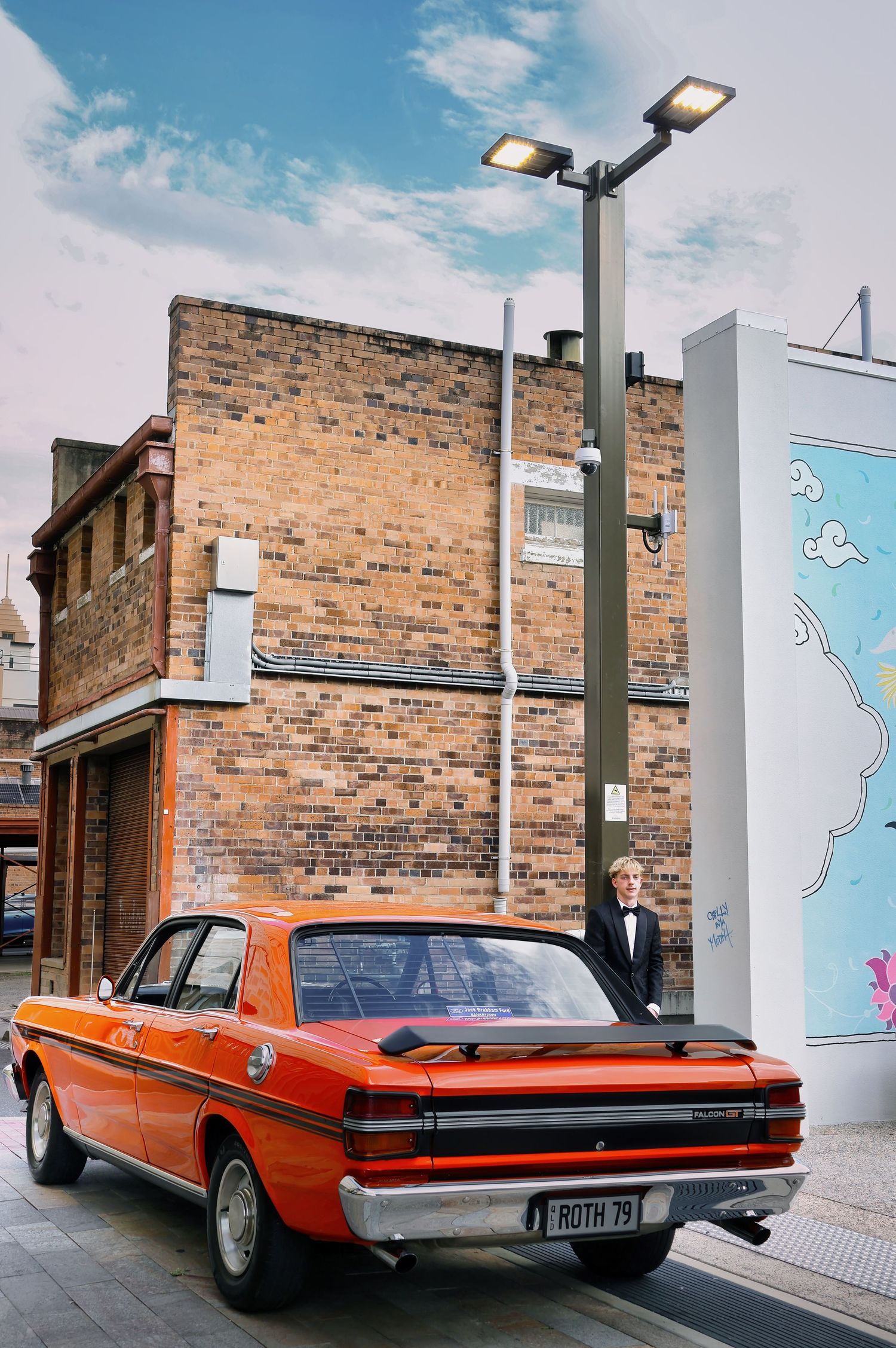 A man in a tuxedo stands beside a vintage orange muscle car on a city street with brick buildings and a mural.