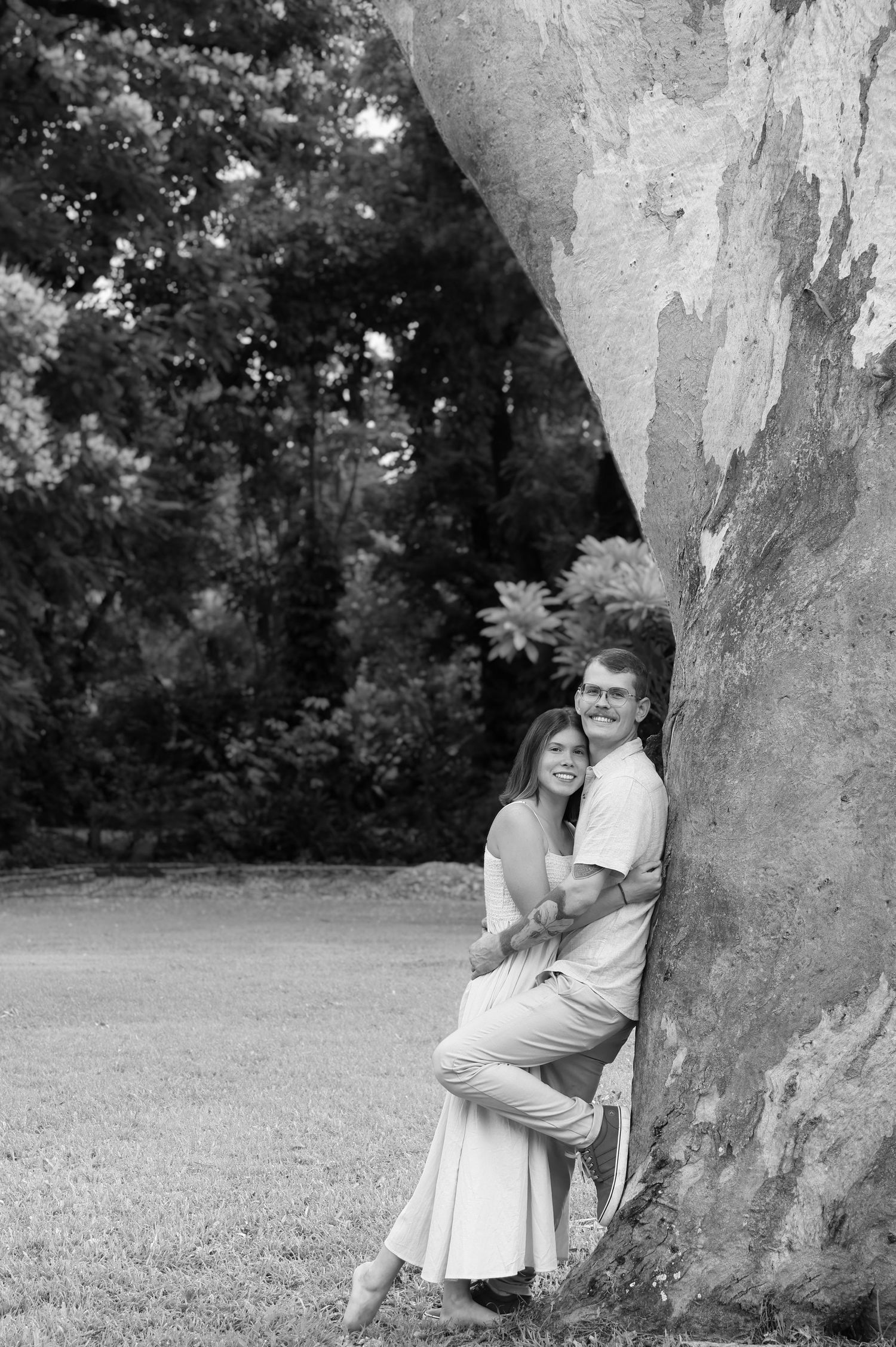 A romantic black and white portrait of a couple embracing against a large tree trunk in a park setting.