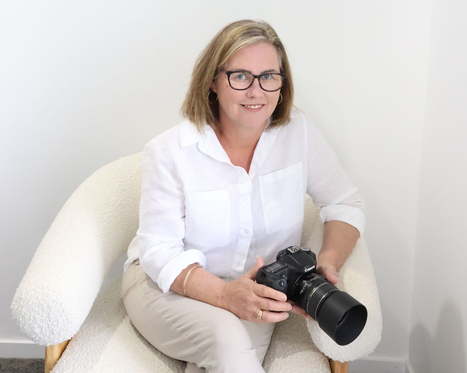 Woman in white shirt holding a DSLR camera, seated in a white chair against a light background.