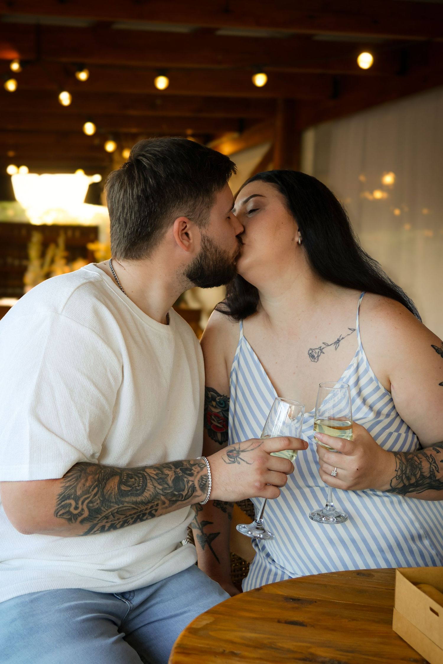 Tattooed couple kissing and clinking wine glasses at a cozy restaurant with warm string lights in background.