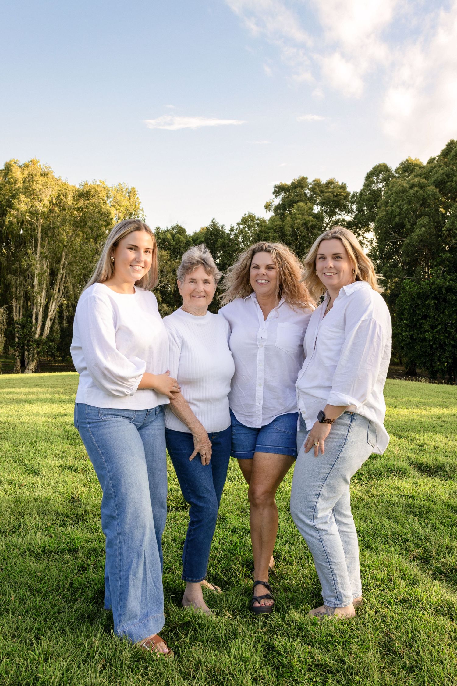 Four women in white tops and jeans smiling together outdoors on green grass with trees and blue sky background.