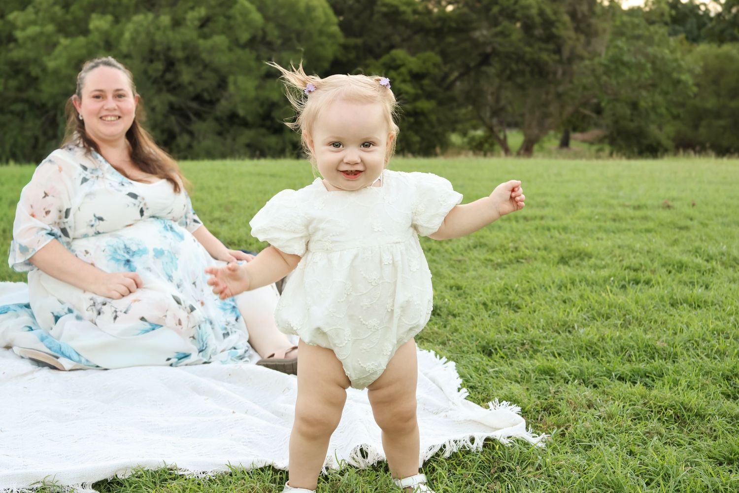 A smiling toddler in a white romper stands on a blanket in a green field while a pregnant woman sits behind her.