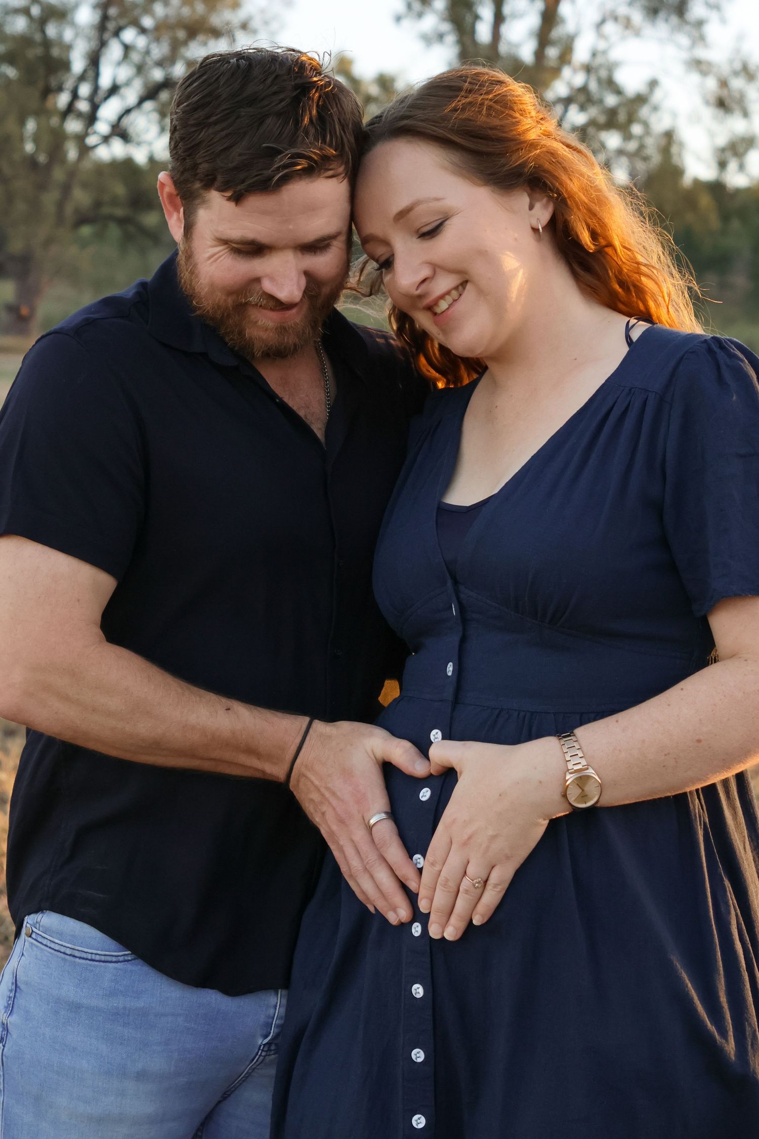 Couple forming a heart shape with hands on pregnant woman's belly outdoors in golden hour light.