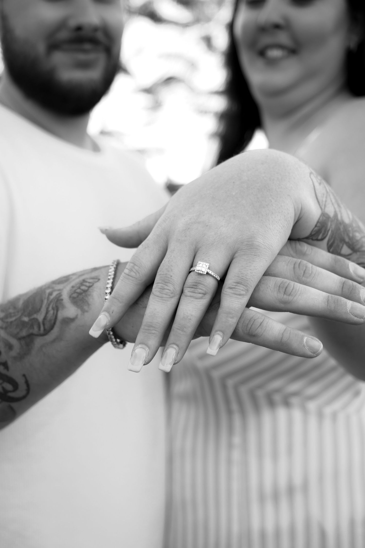 Black and white photo of a couple showing off an engagement ring with diamond solitaire, both with tattooed arms.