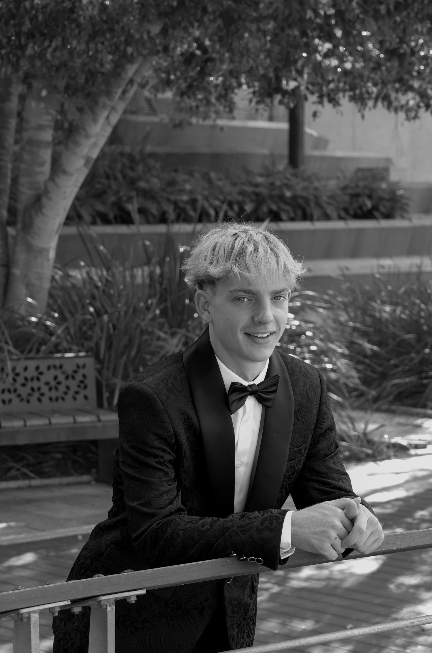 Young man in black tuxedo and bow tie smiling outdoors, leaning on railing in a black and white portrait photo.