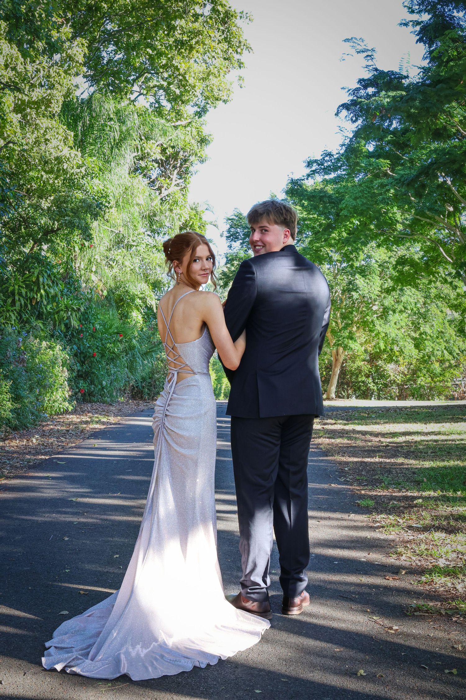A couple in formal attire poses on a sunlit path, woman in backless white gown, man in black suit, surrounded by lush trees.