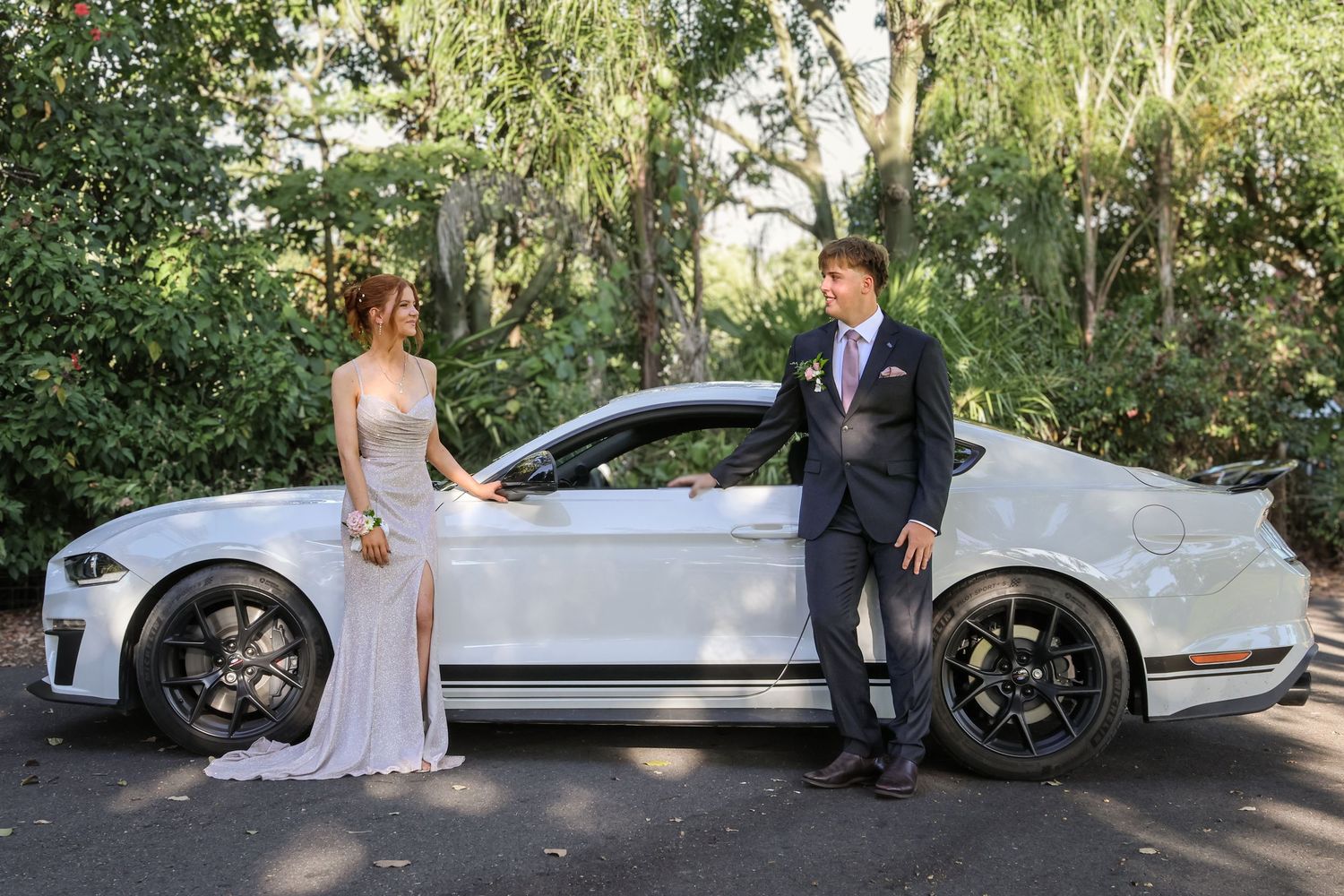 Elegant couple in formal attire posing beside a white Ford Mustang for a prom or wedding photo.