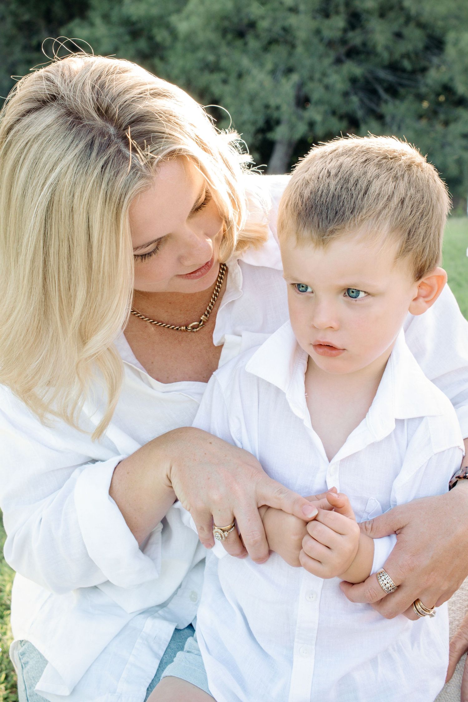 Blonde mother in white shirt tenderly holding young boy outdoors in green park setting.
