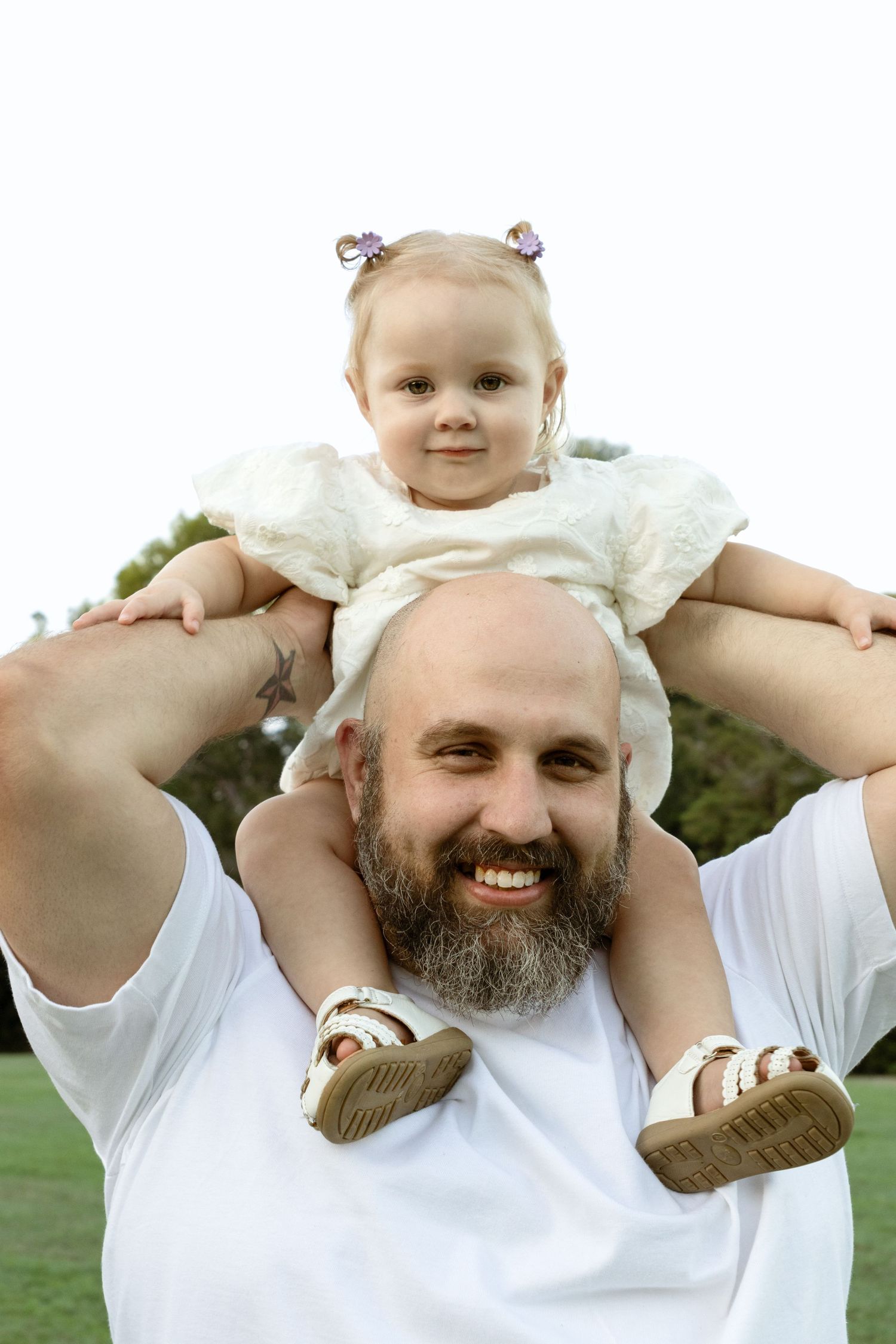 Smiling bald bearded man holding a cute toddler girl on his shoulders outdoors in a park.