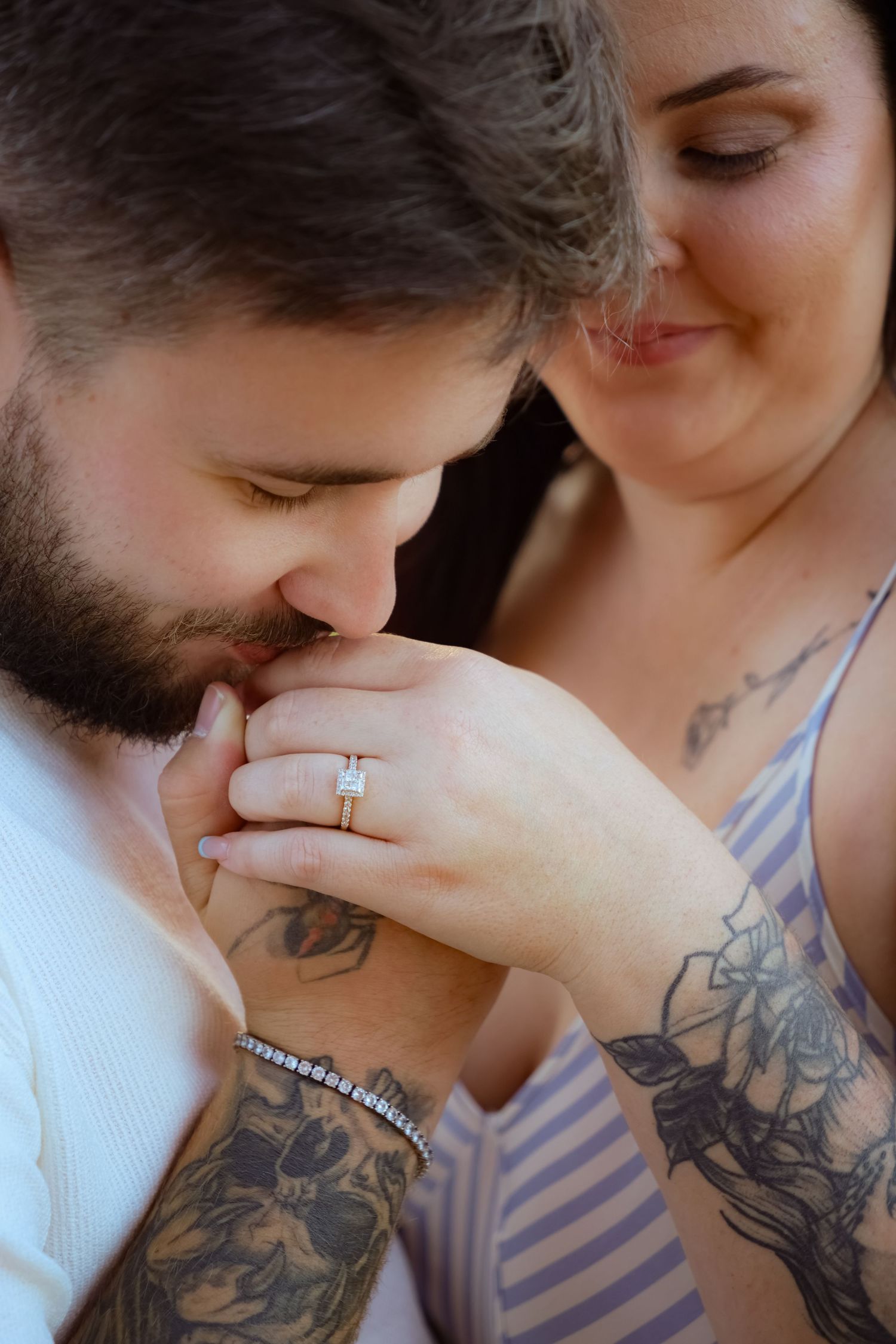 Bearded tattooed man kissing hand of smiling woman wearing diamond engagement ring, romantic proposal moment.