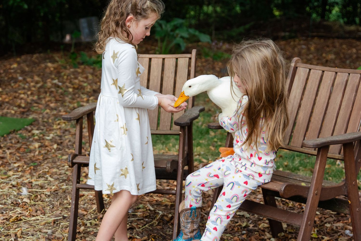 Two children in white and floral outfits sit on wooden chairs in a garden setting sharing snacks together at Delafield home session