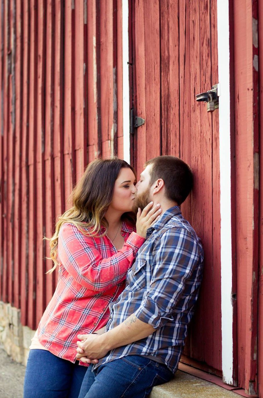 A romantic couple in plaid shirts share a tender moment against a rustic red barn wall.