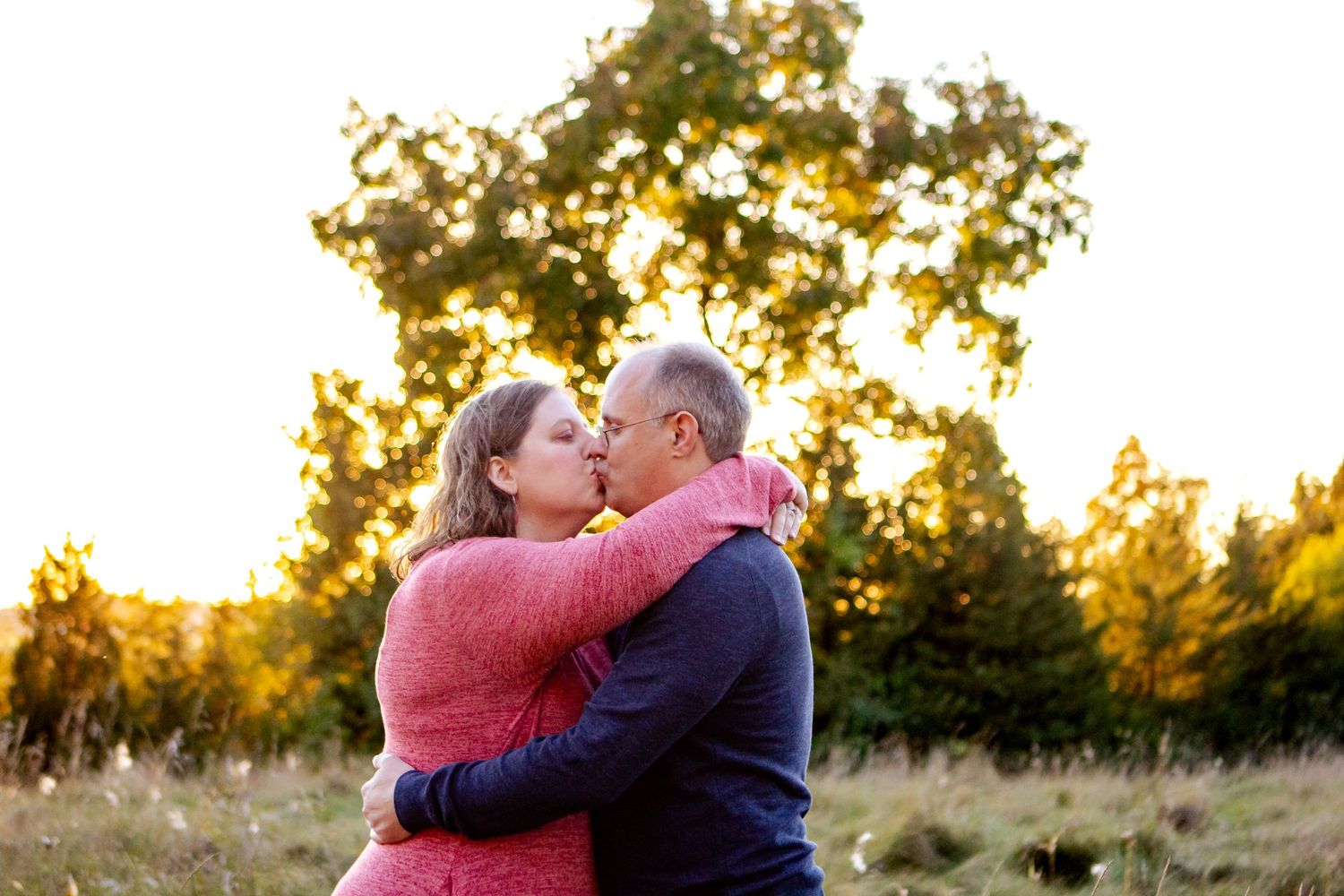 Couple embraces in a golden sunset field with autumn trees and glowing light in the background at an Oconomowoc family session