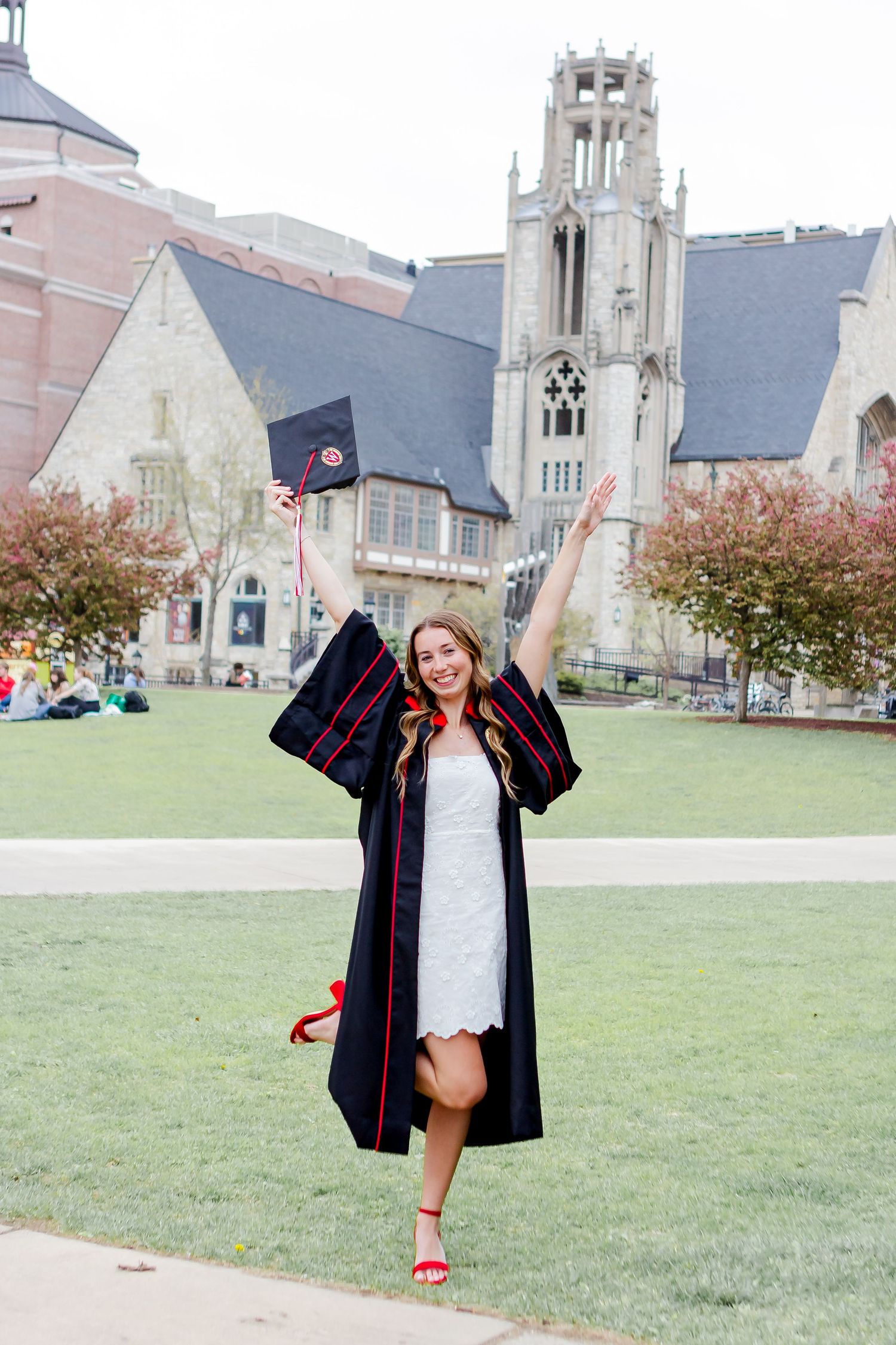 Graduate celebrates on campus lawn with historic university buildings in background.