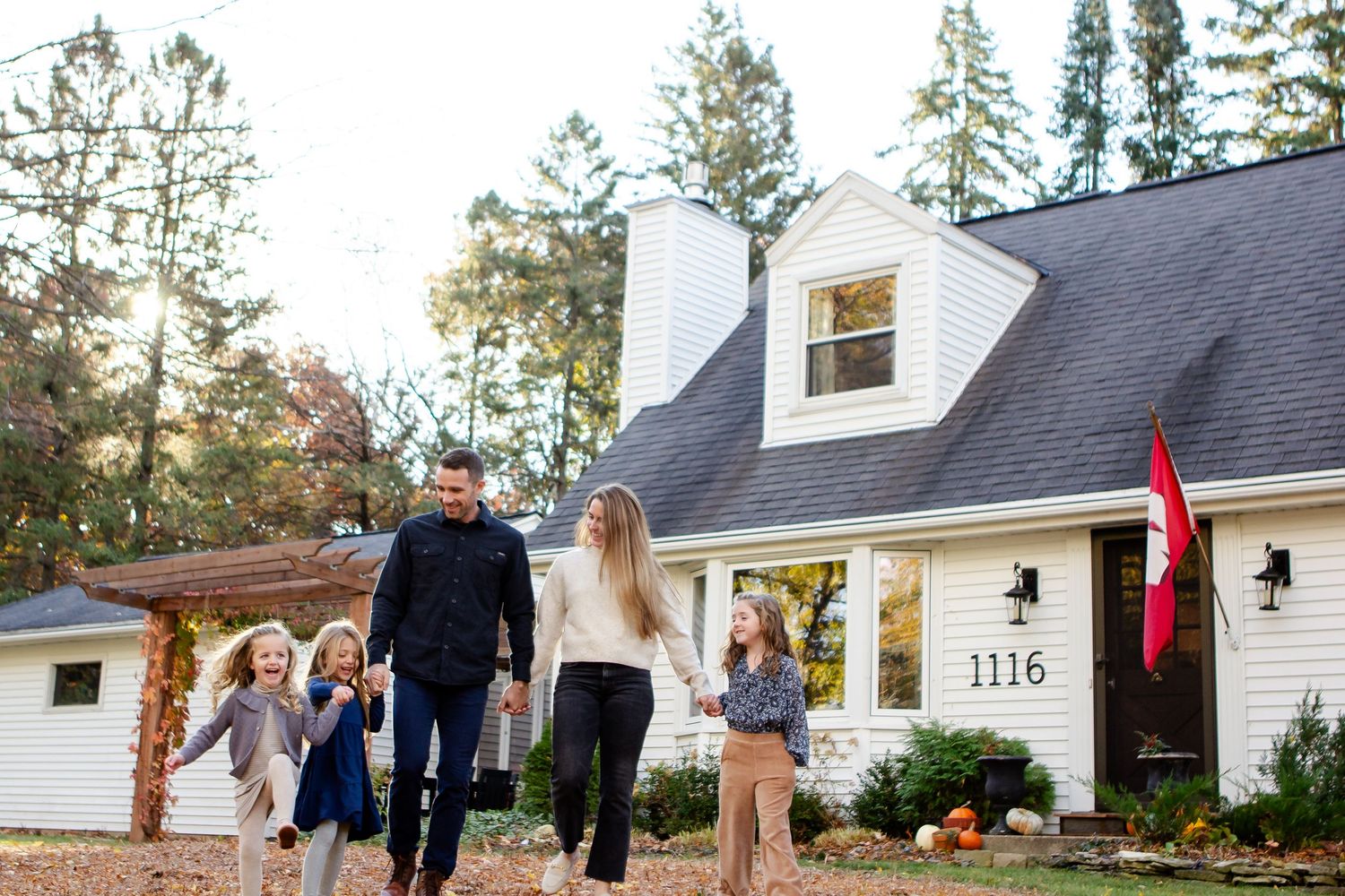 A family walks together outside their white Cape Cod style home with a dormer window and Canadian flag at sunset during a Delafield session