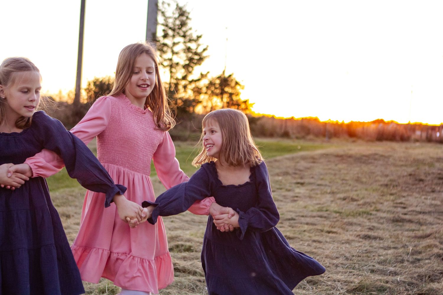 Three people in flowing dresses dance together in a field at sunset with golden light behind them at an Oconomowoc family session