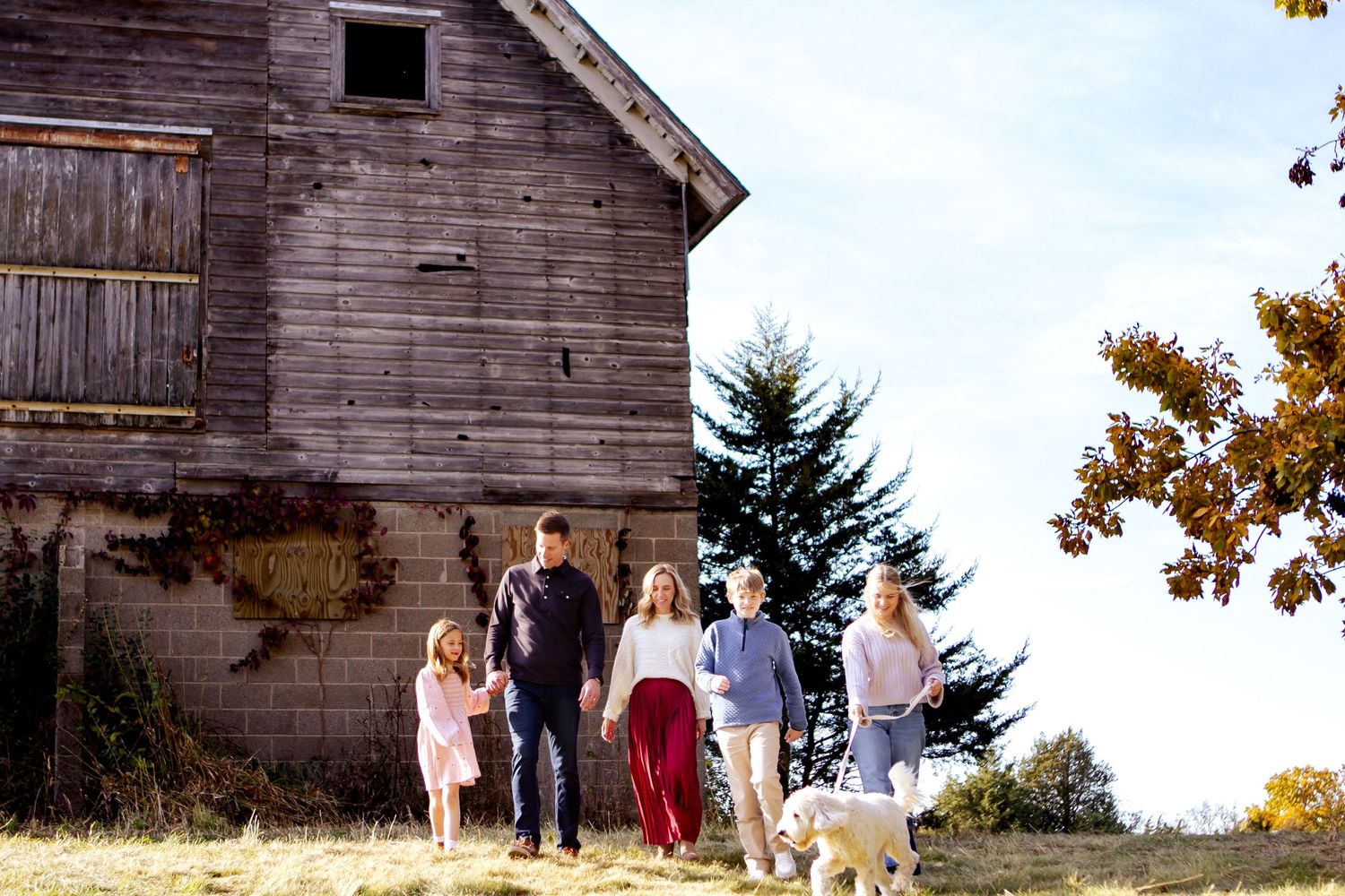 A group walks together with their dog near a rustic wooden barn on an autumn hillside at a Lake Country Family session