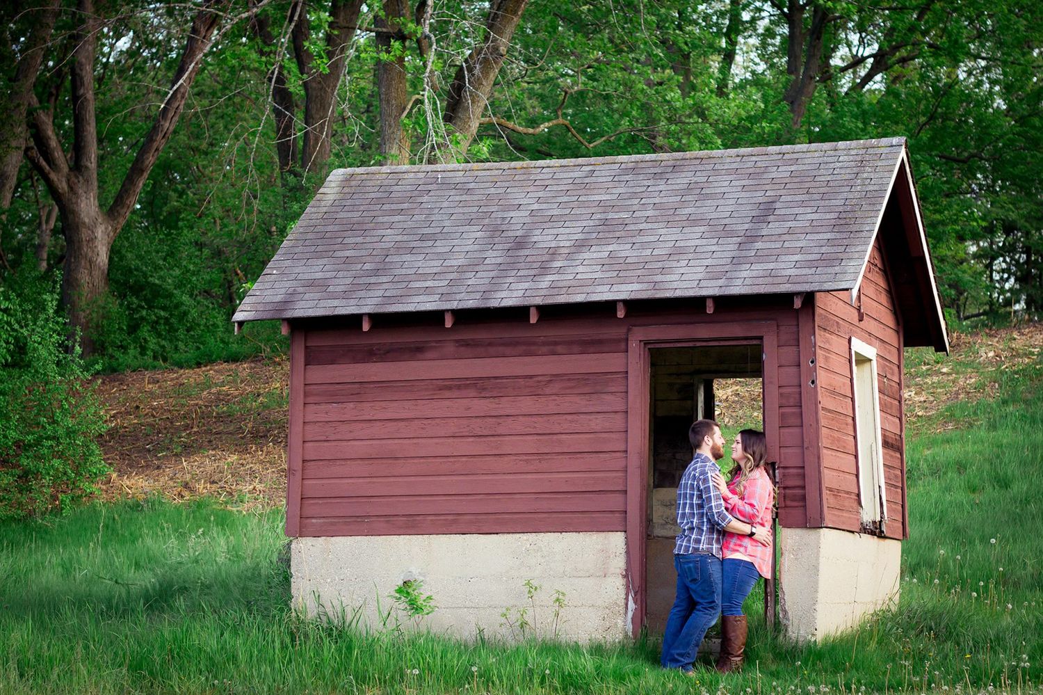 Small red wooden shed with gray shingle roof sits nestled among green trees on grassy hillside.