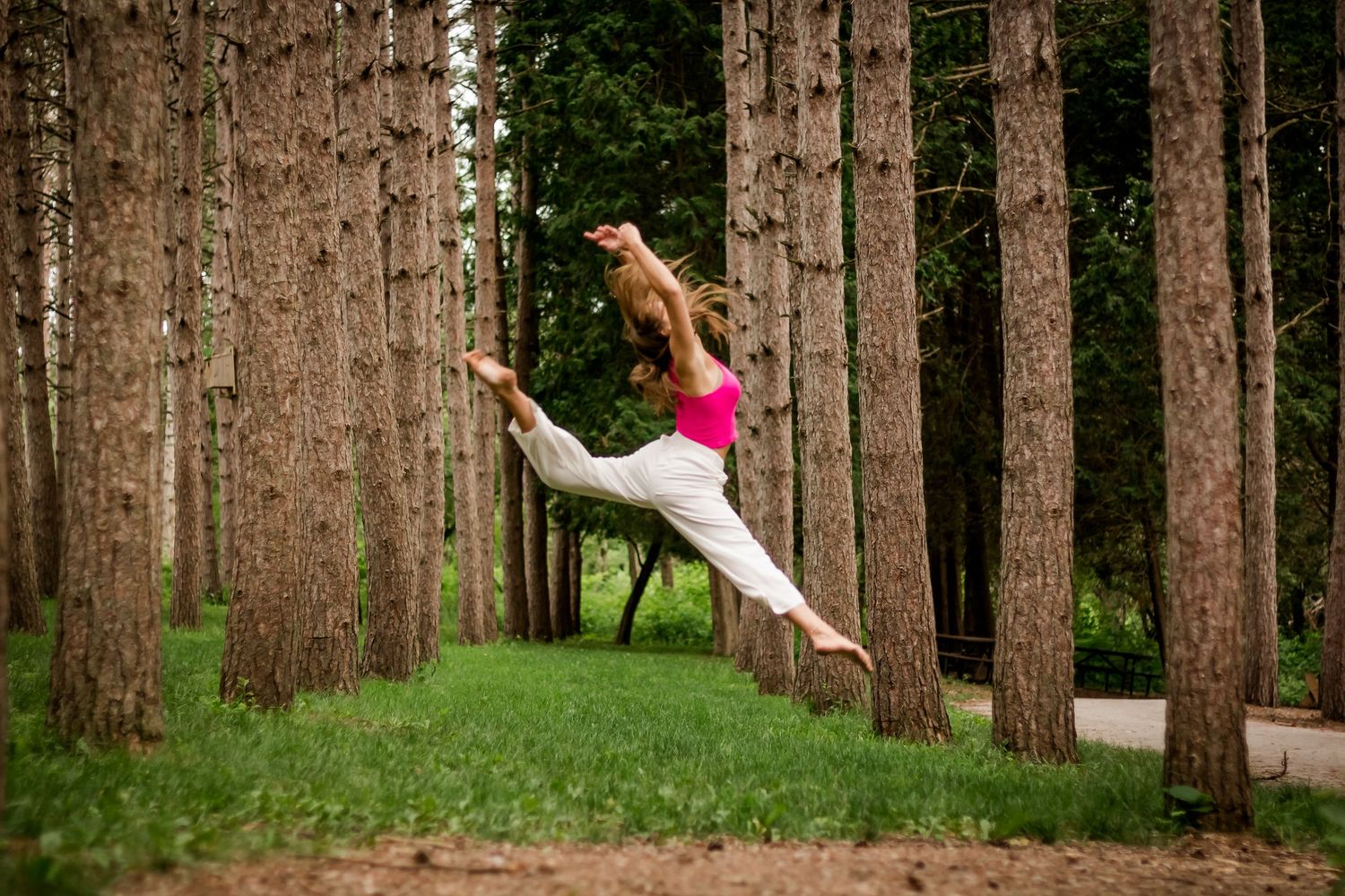 A dancer performs a graceful aerial split jump between tall pine trees in a forest setting at Retzer Nature Center in Waukesha