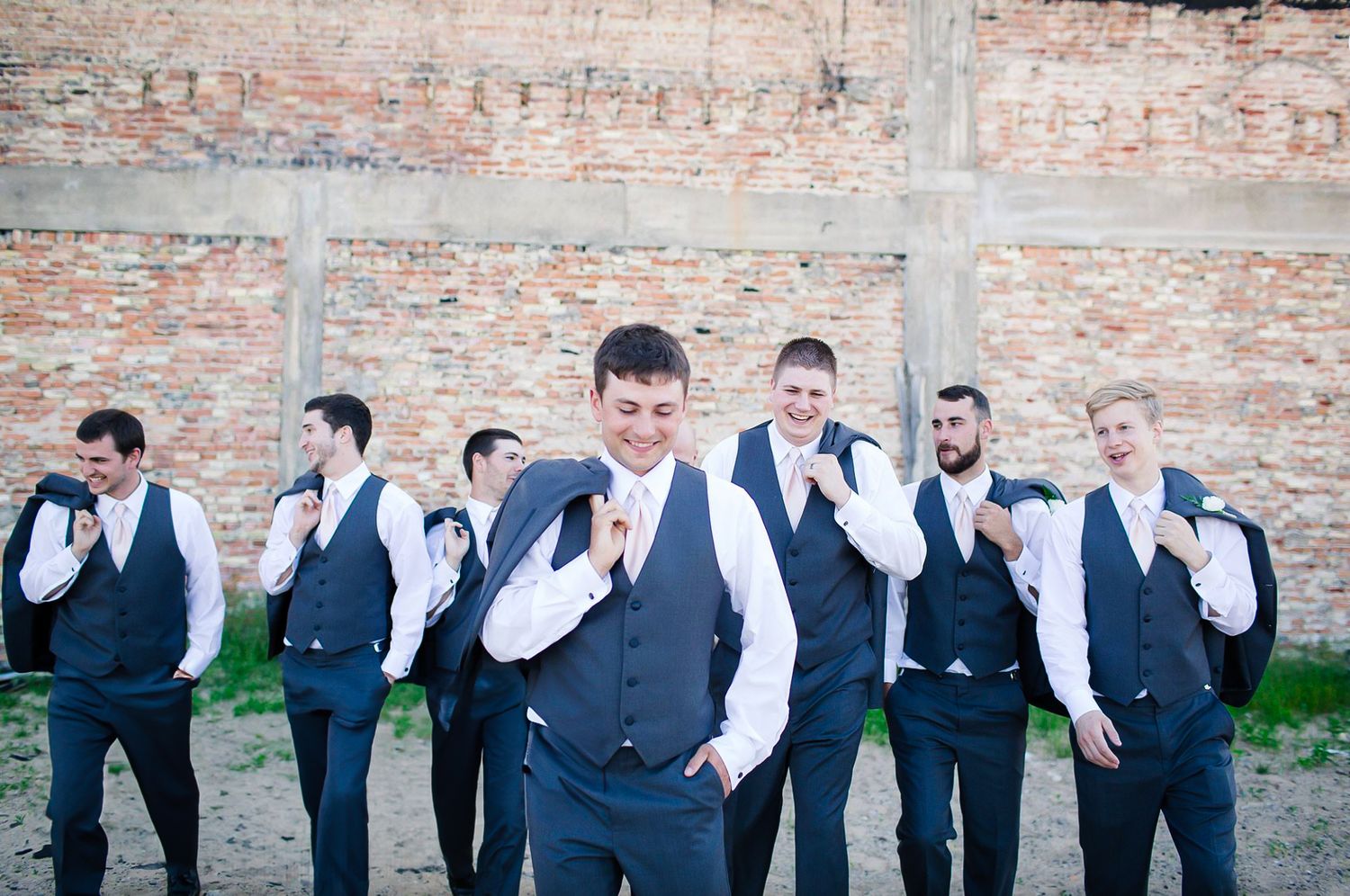 Wedding party in navy suits and vests poses against rustic brick wall backdrop during formal photo session in Milwaukee