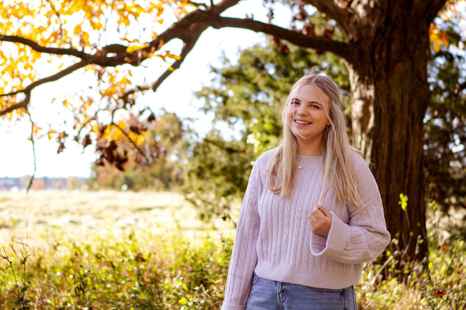 A woman in a cozy pink sweater sits outdoors among autumn foliage and golden sunlight at a Lake Country Family session