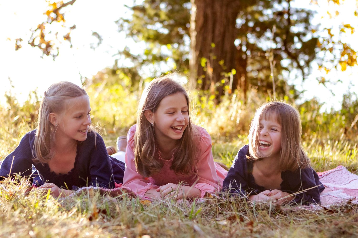 Three young sisters lying on a pink blanket in sunny grass laughing together on a warm autumn day at an Oconomowoc family session