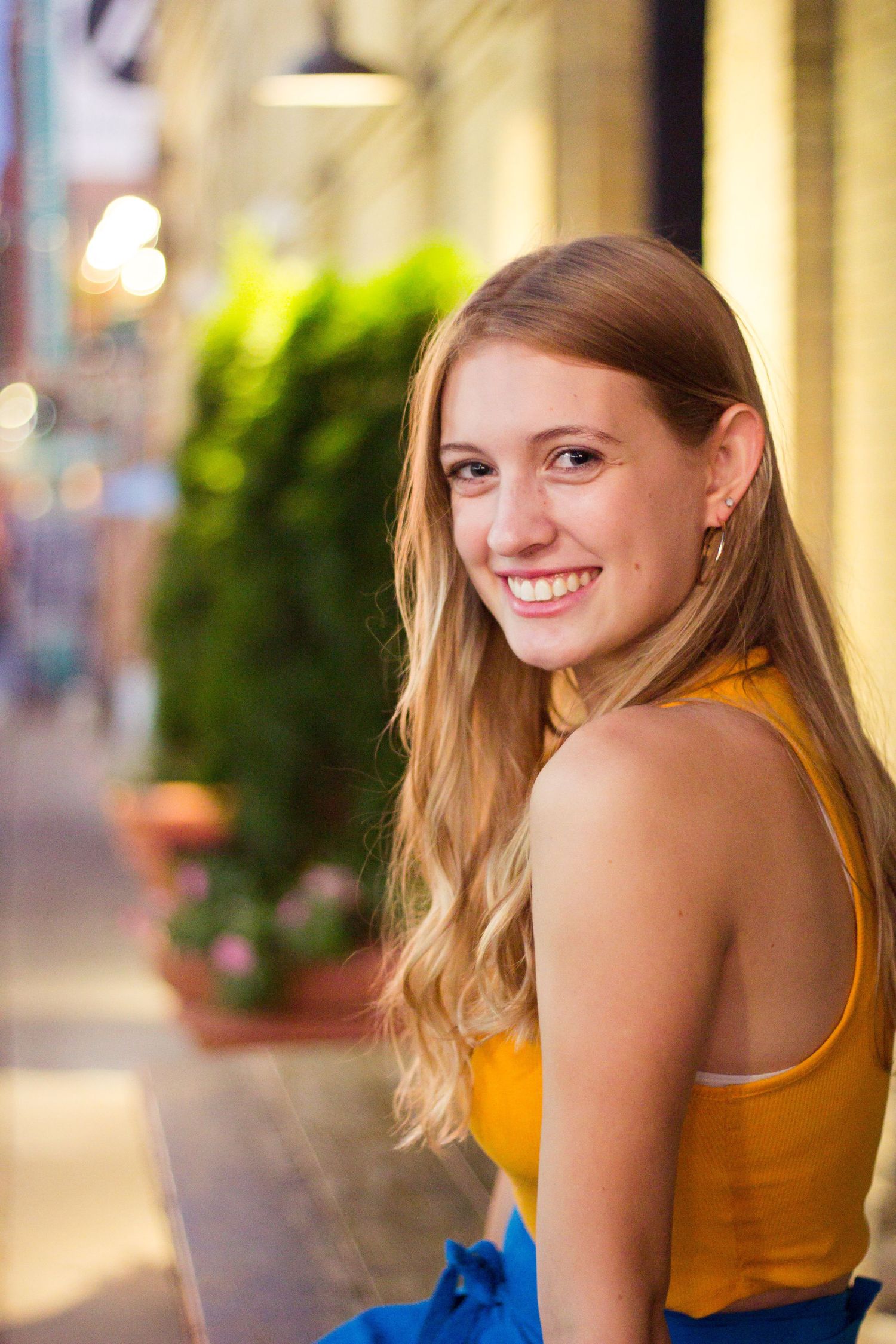 Woman in yellow tank top smiling in outdoor setting with warm lighting and urban backdrop.