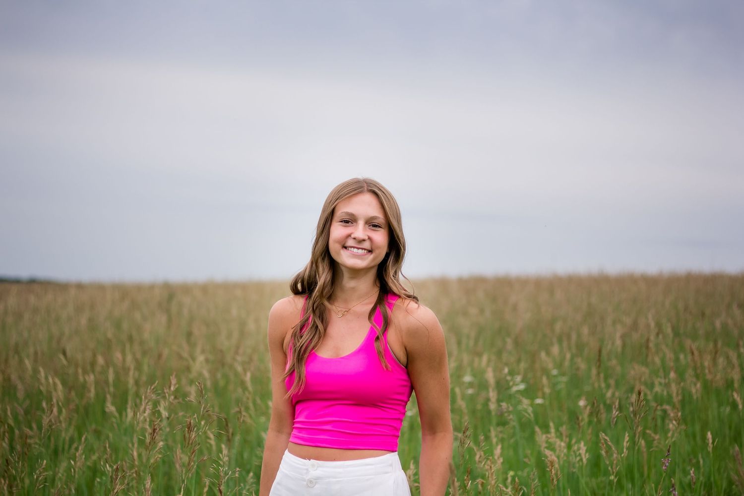 Woman wearing bright pink tank top stands smiling in tall grass field under cloudy sky.