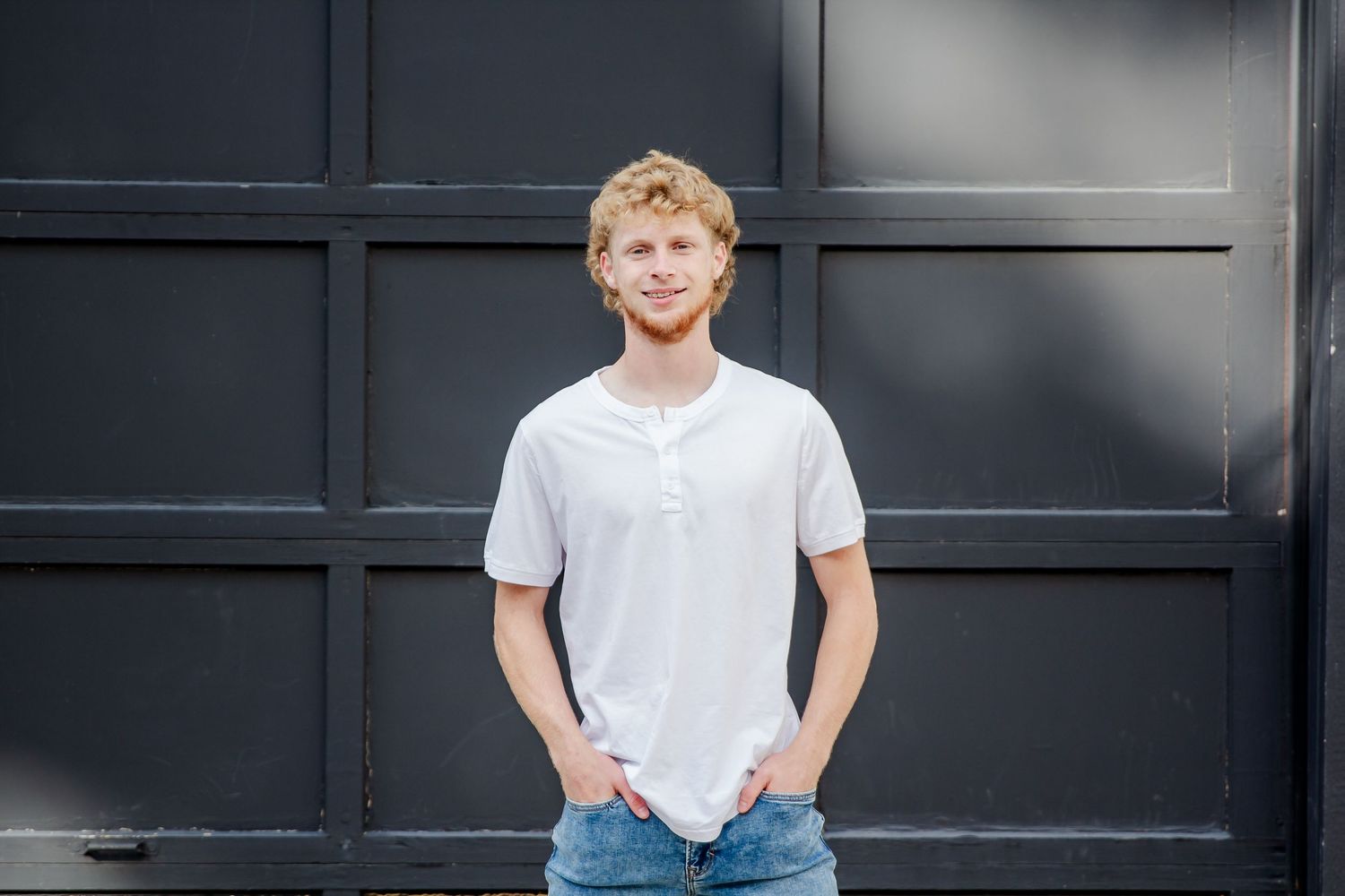 A casual portrait against a dark wall wearing a white henley shirt and jeans with hands in pockets.