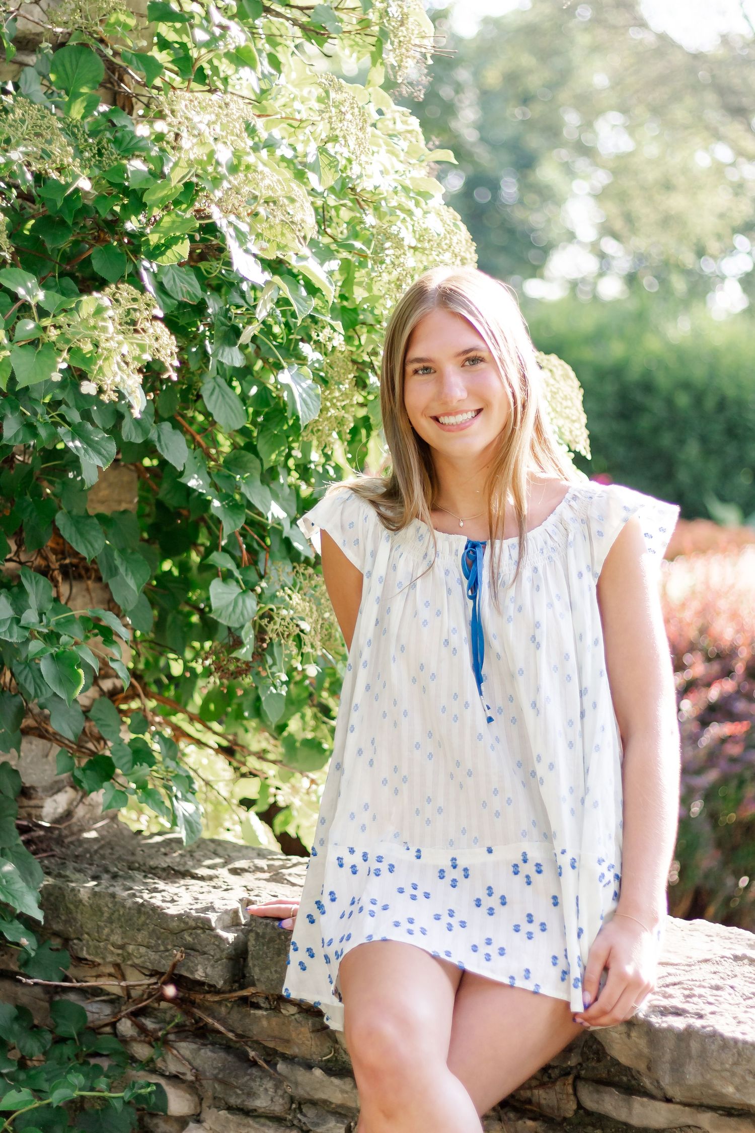 A summer portrait in a white eyelet dress with blue accents beside flowering vines.