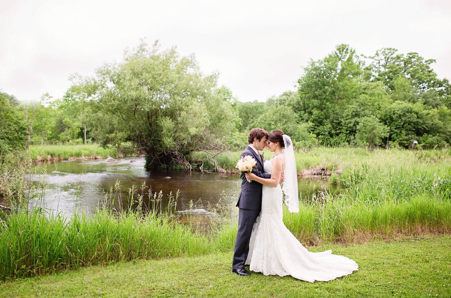 Wedding couple posing for romantic photos by a serene waterfront with natural greenery.