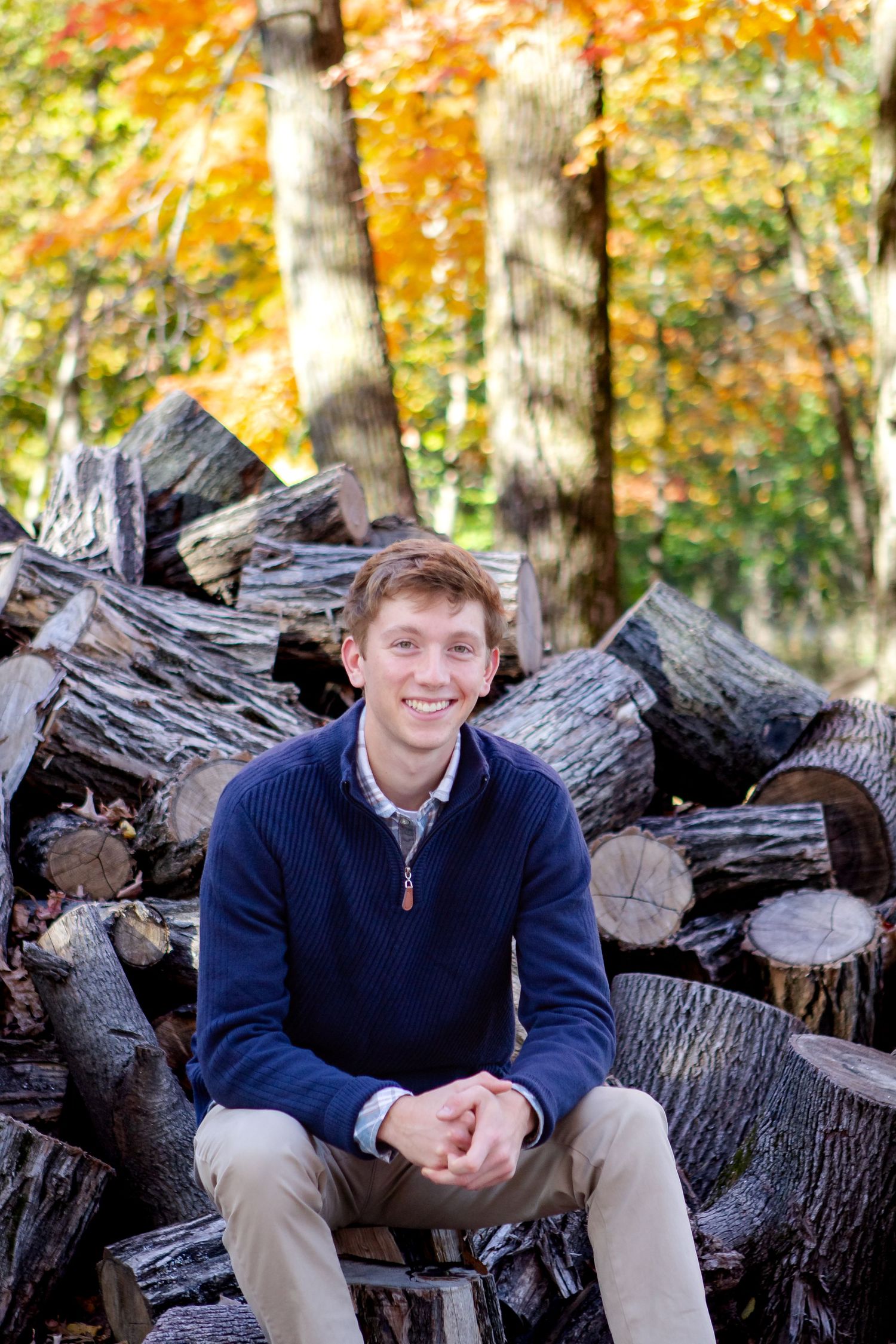 Four photos show someone sitting on a woodpile surrounded by autumn foliage in a casual outdoor setting during his Hartland session
