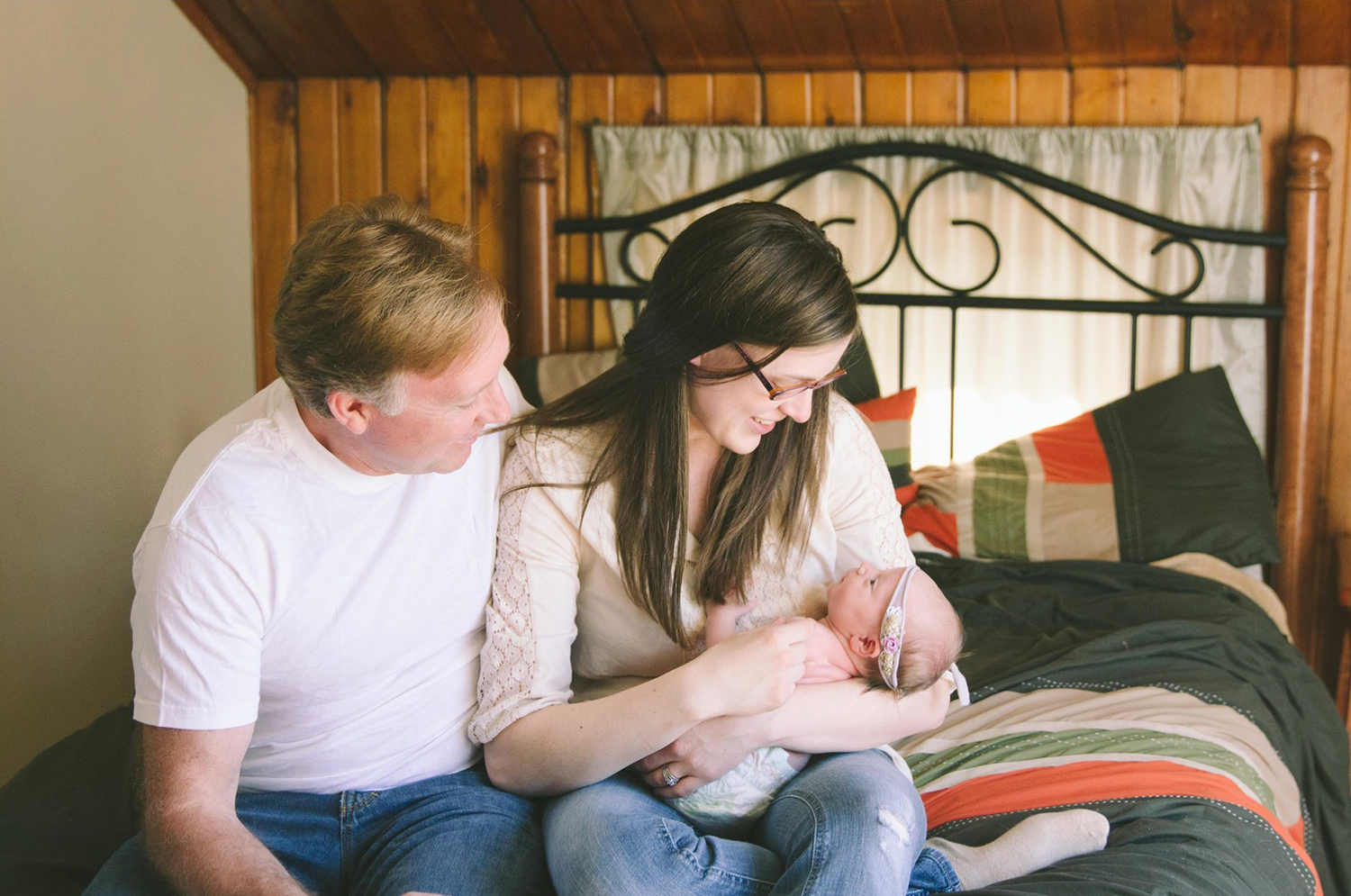 A couple sits together on a bed with patterned bedding in a room with wooden paneled walls.