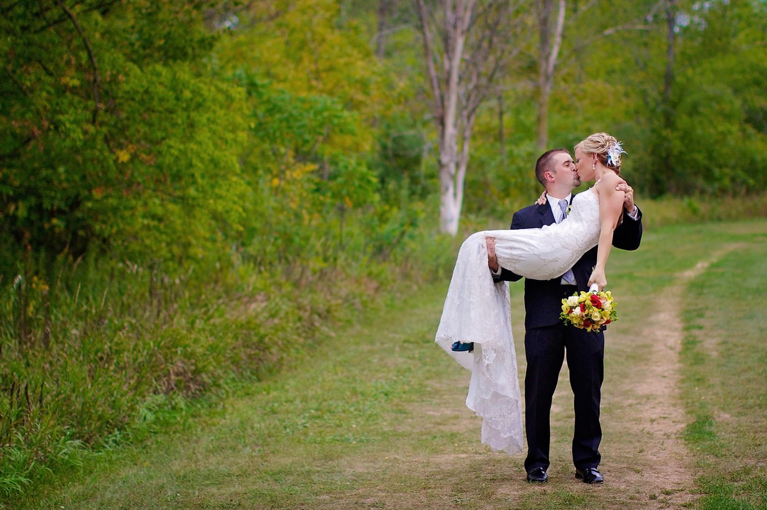 A couple shares a romantic moment on a grassy path surrounded by lush green trees.