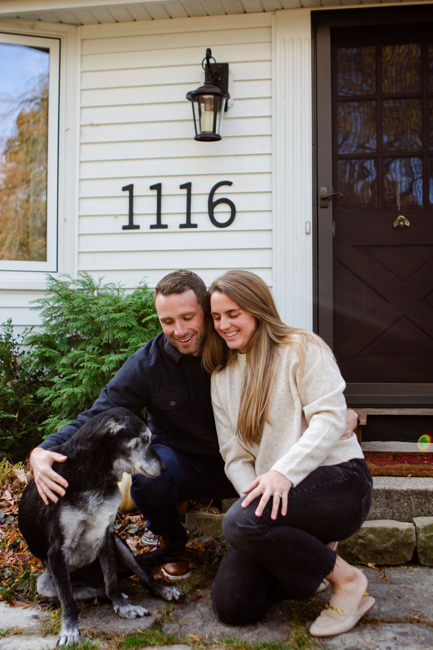 A couple poses with their black dog in front of a house entrance with the number 1116.