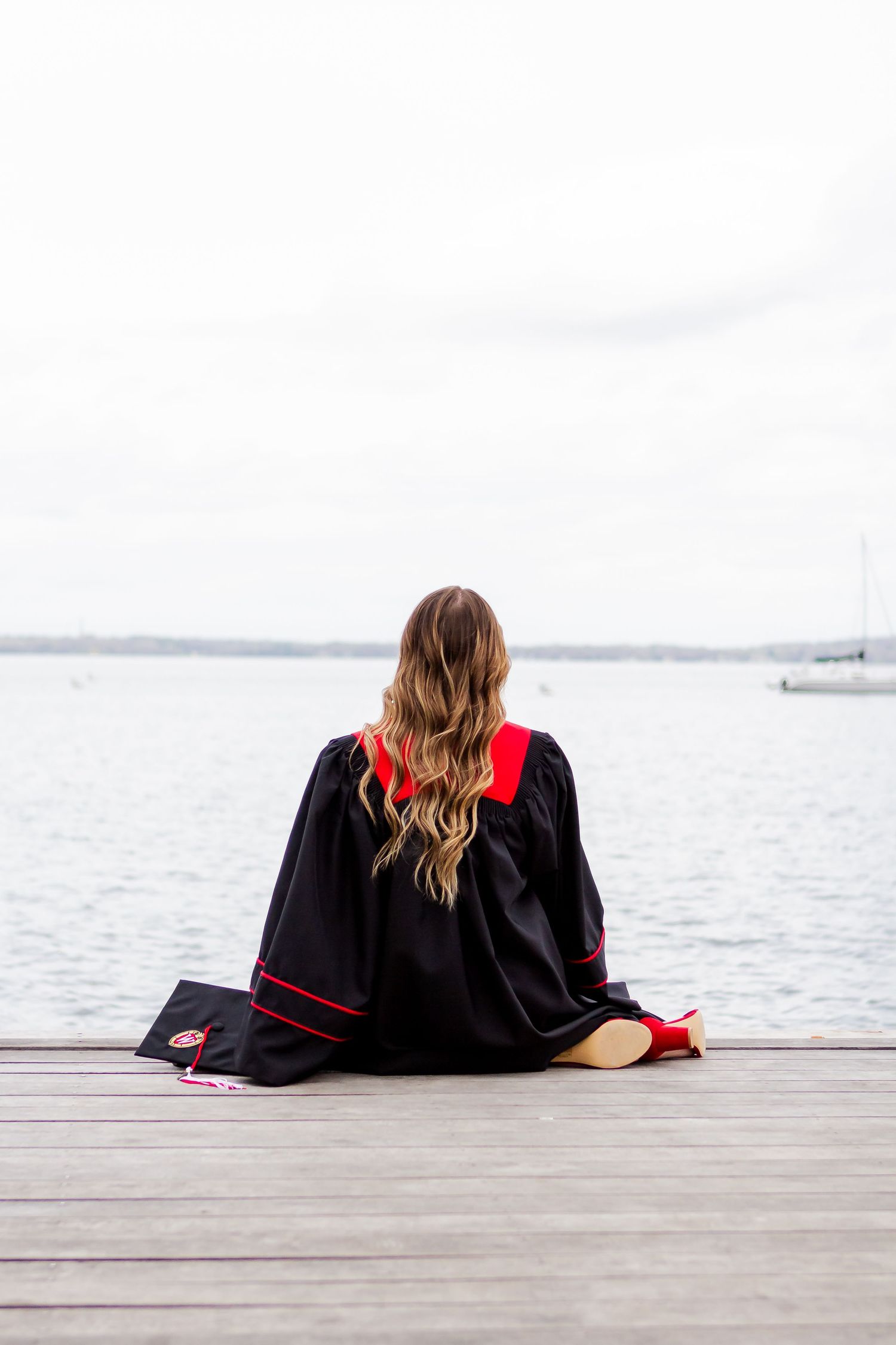 Graduate in black and red regalia sitting on a wooden dock overlooking a serene lake.