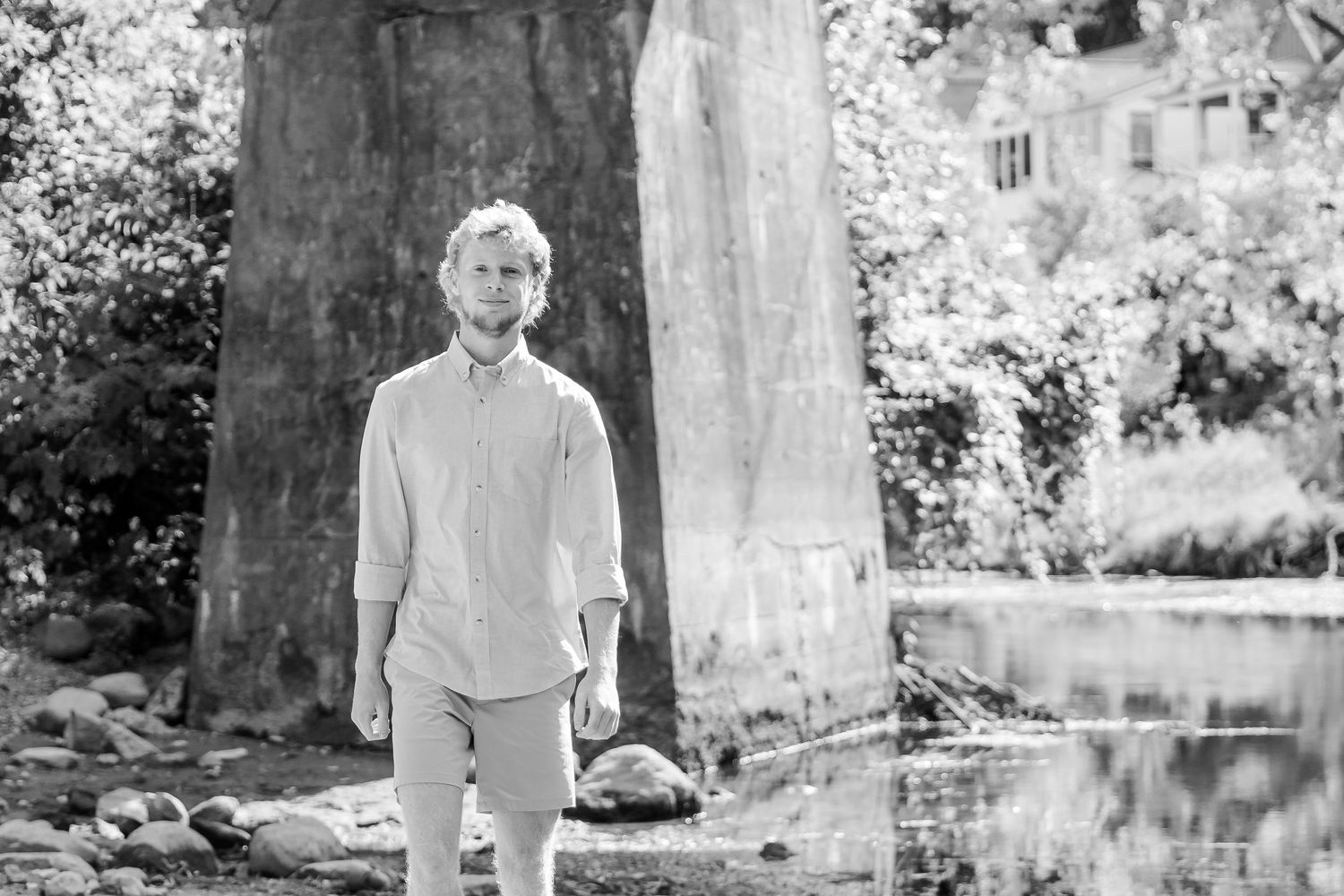 A person stands near a large tree trunk beside a reflective pond in a black and white photograph during a Michigan senior session