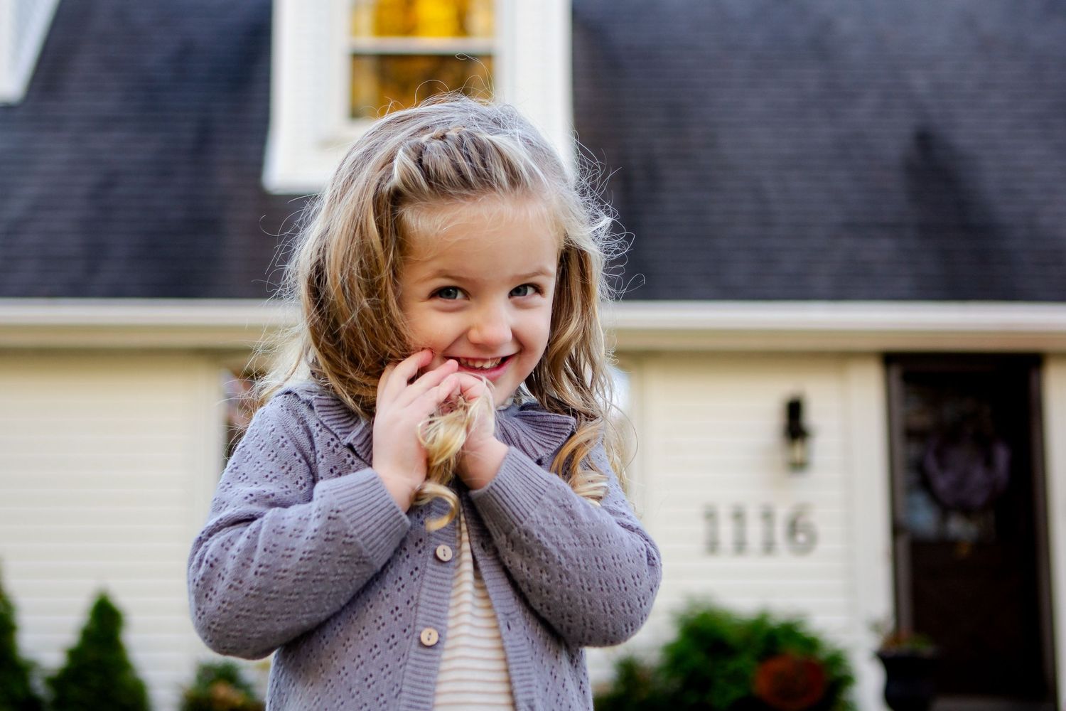 Young child in gray sweater stands outside house with hands covering face in playful expression during a Delafield photo shoot