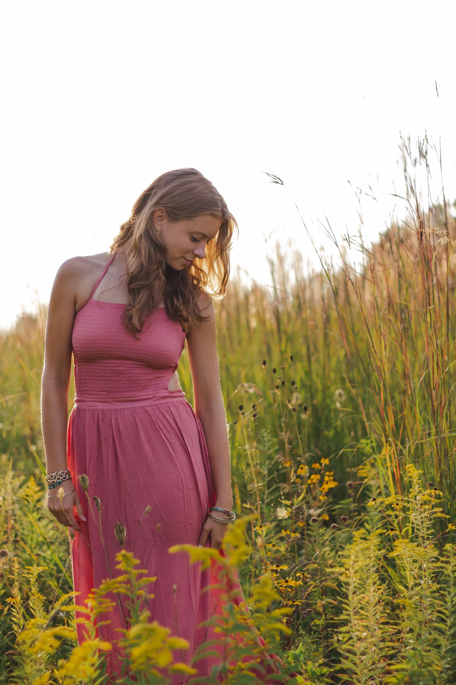 Person in pink dress walking through field of golden wildflowers at sunset.