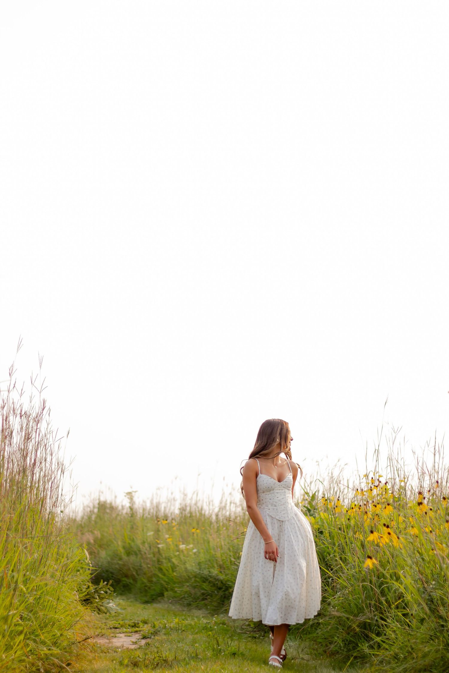 A person in a white dress walks through a field of yellow wildflowers and tall grass at sunset during a session at Retzer Nature Center