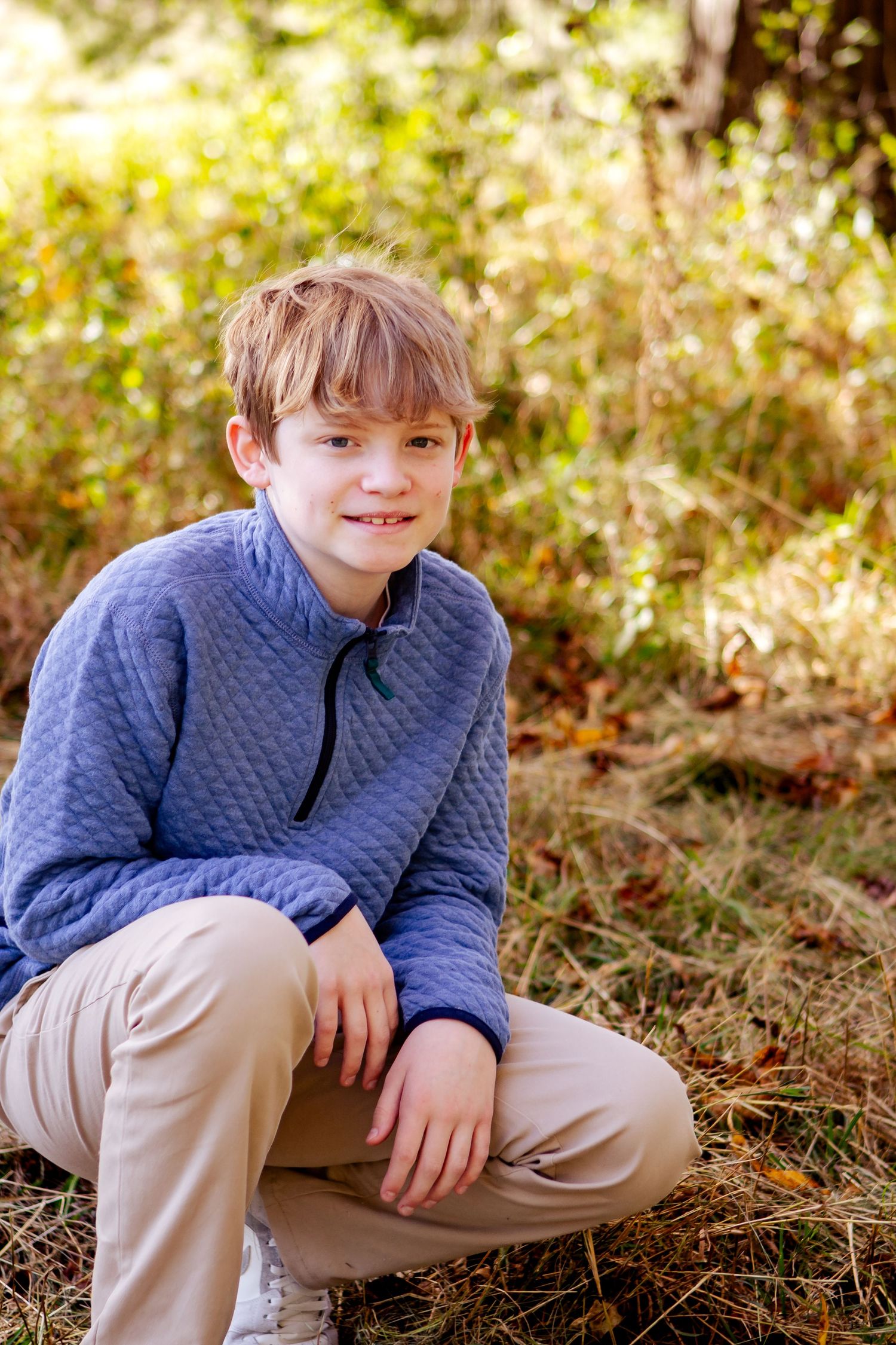 A young person in a blue quilted pullover and khaki pants crouches outdoors in an autumn setting with golden bokeh background in Oconomowoc