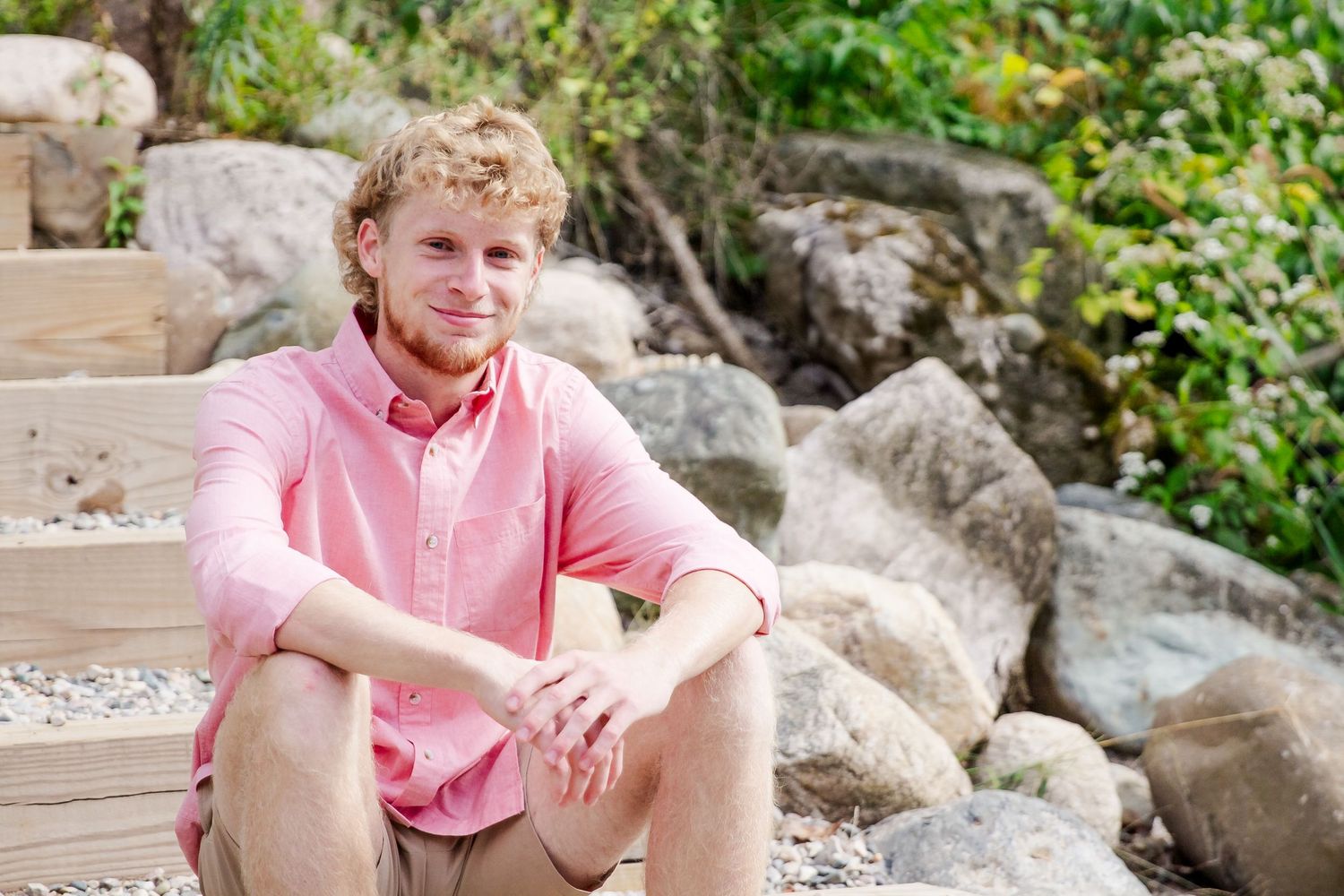 Person in a coral pink shirt sits casually outdoors among rocks and greenery in a natural setting during a Michigan senior session