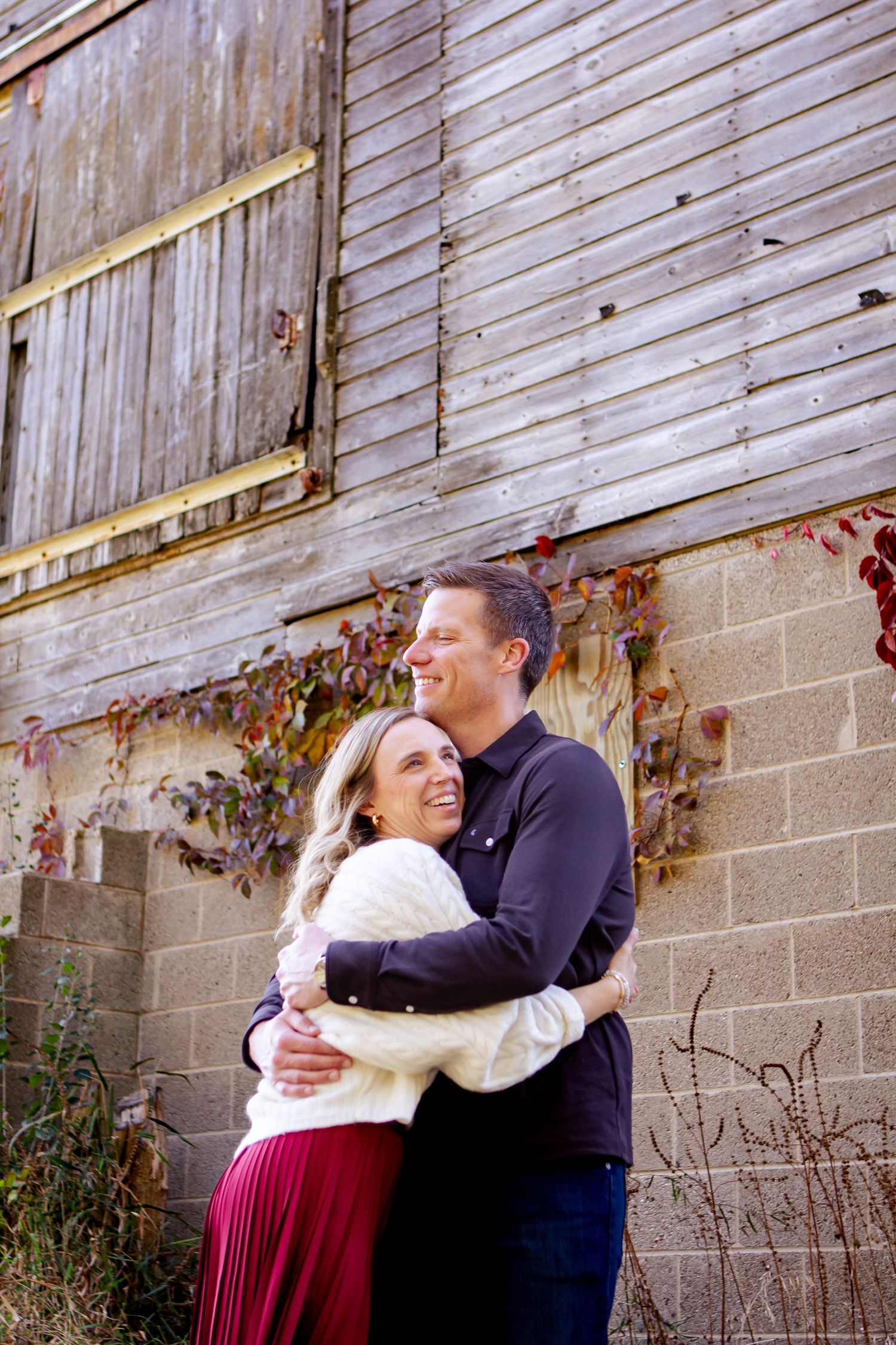 A couple embraces and shares a laugh in front of a rustic wooden wall and concrete blocks at an Oconomowoc family session