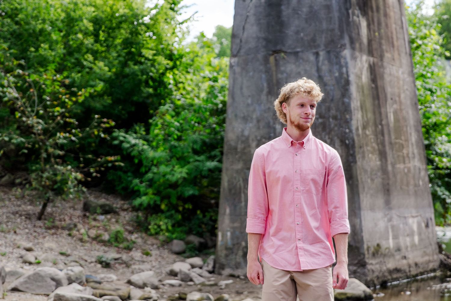 A person in a pink button-down shirt and khaki pants stands in front of a large concrete pillar surrounded by greenery at Michigan session