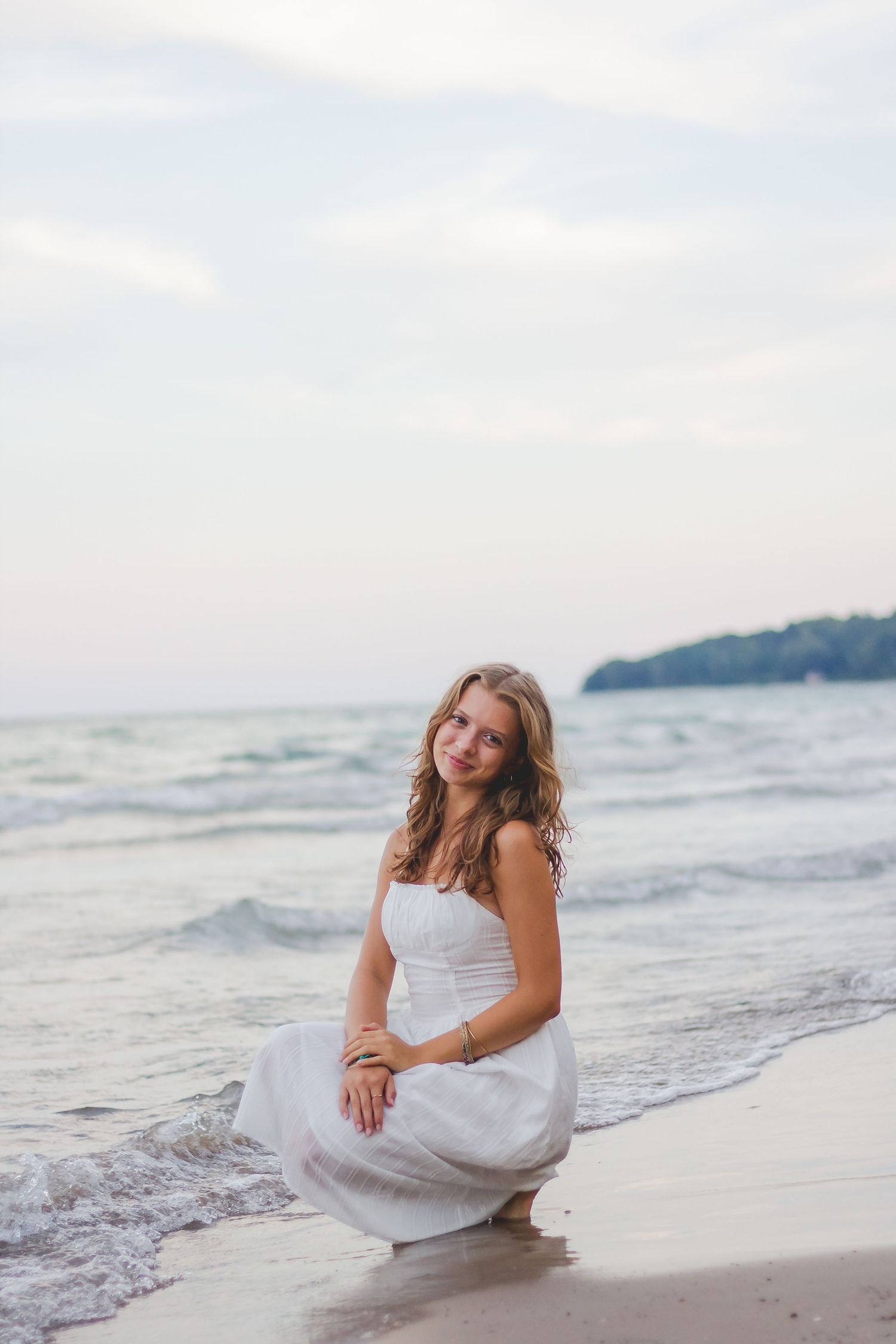 Someone in a white dress sits gracefully on a sandy beach with waves in the background.