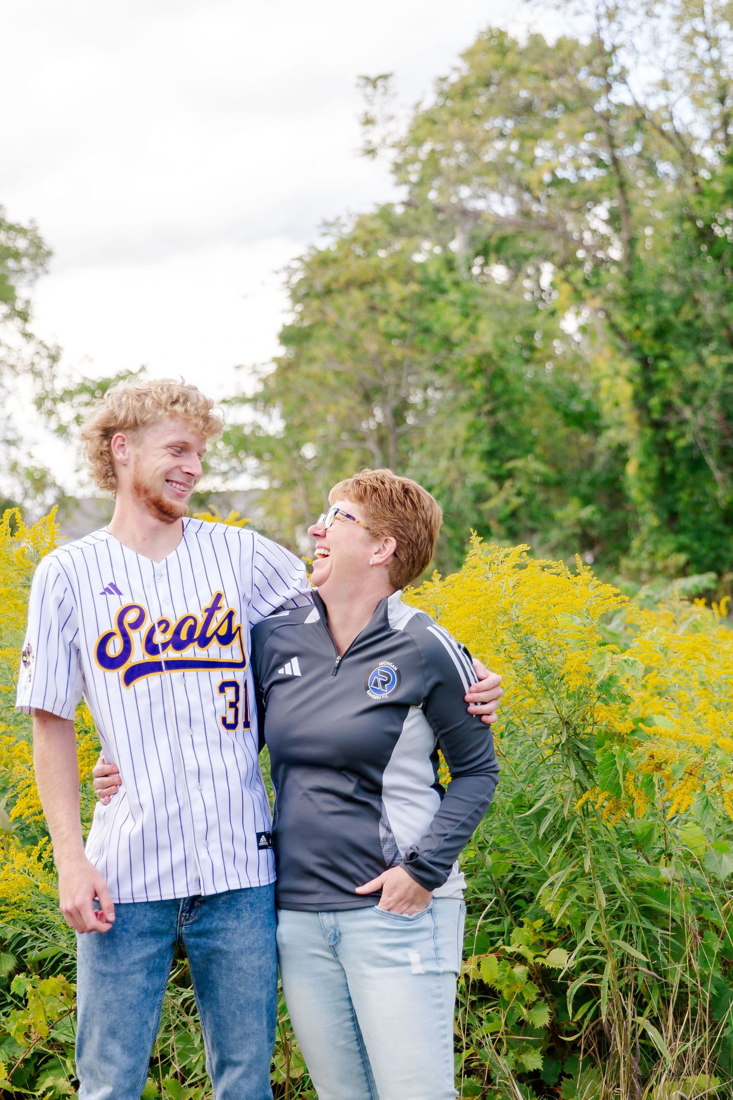 A mom and son share joyful laughter together in a field of blooming yellow goldenrod on a sunny autumn day during a Michigan senior session