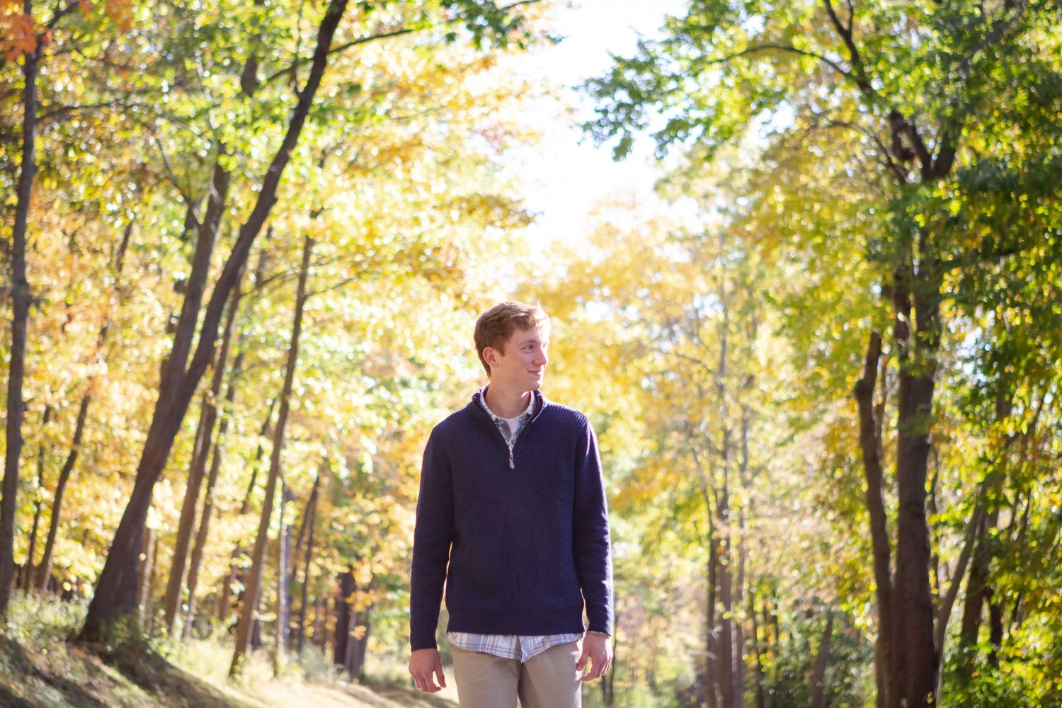 A boy walks down a tree-lined path with golden autumn foliage overhead on a sunny afternoon during his Hartland senior session