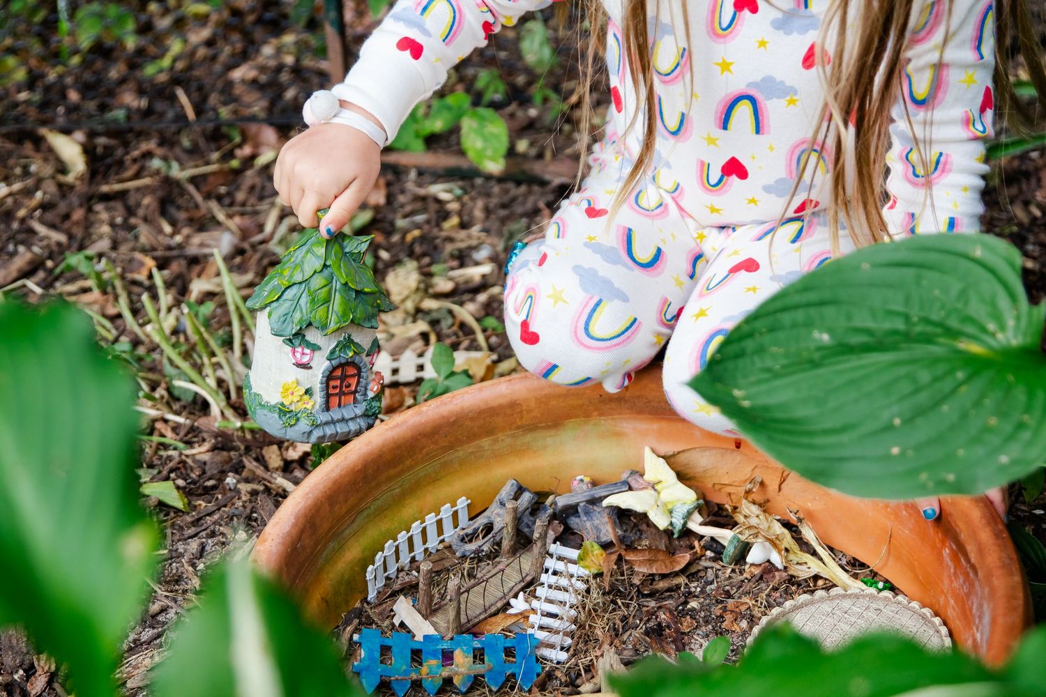 A child wearing rainbow print clothing plays with toy trains in a garden with hosta plants and terracotta pots at Delafield home session